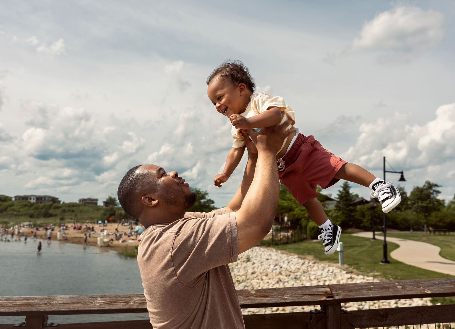 Father joyfully lifting smiling baby in the air outdoors near a lake on a cloudy day.
