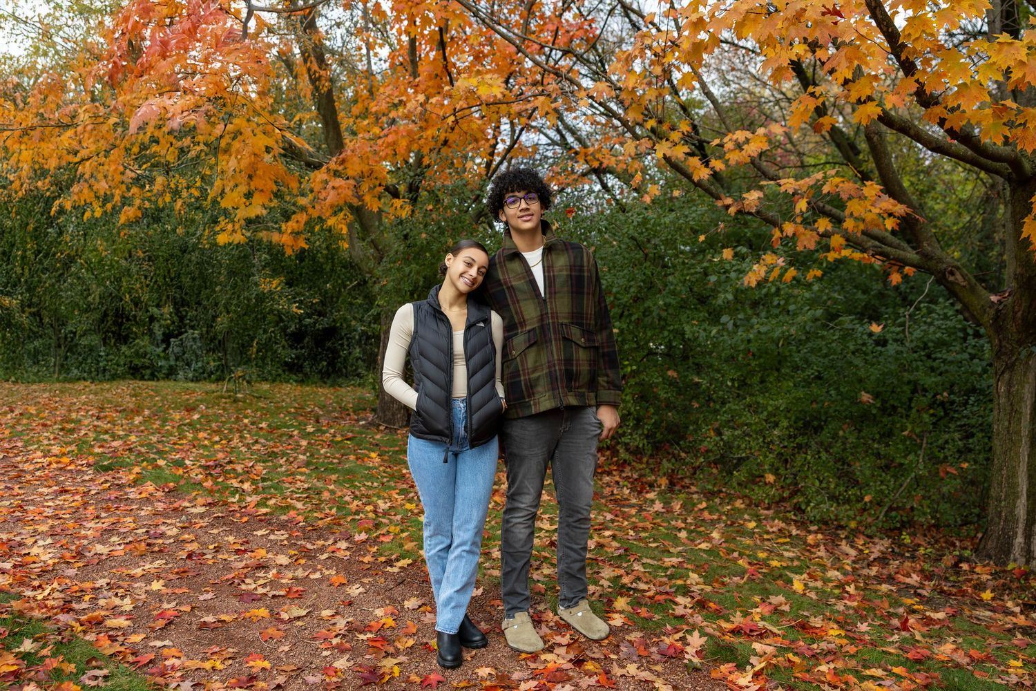 A smiling couple stands together in a scenic autumn park, surrounded by colorful fallen leaves and orange foliage.