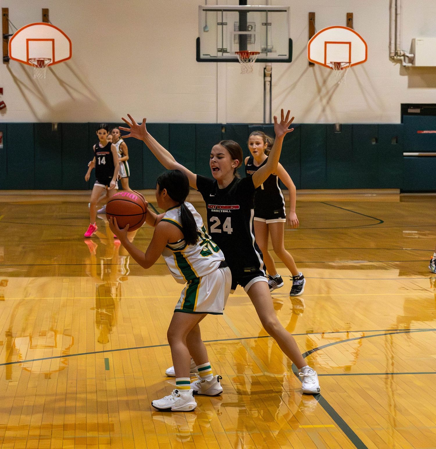 Girls basketball game with player #24 defending opponent holding ball in indoor gymnasium with hardwood floors.