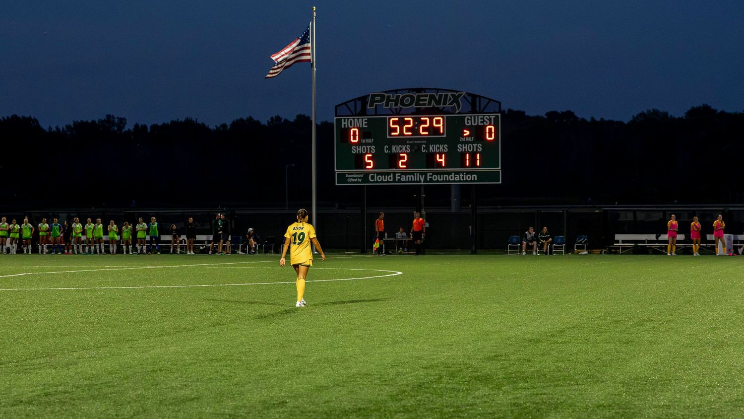 Soccer field at dusk with scoreboard glowing and American flag flying against dark blue sky.