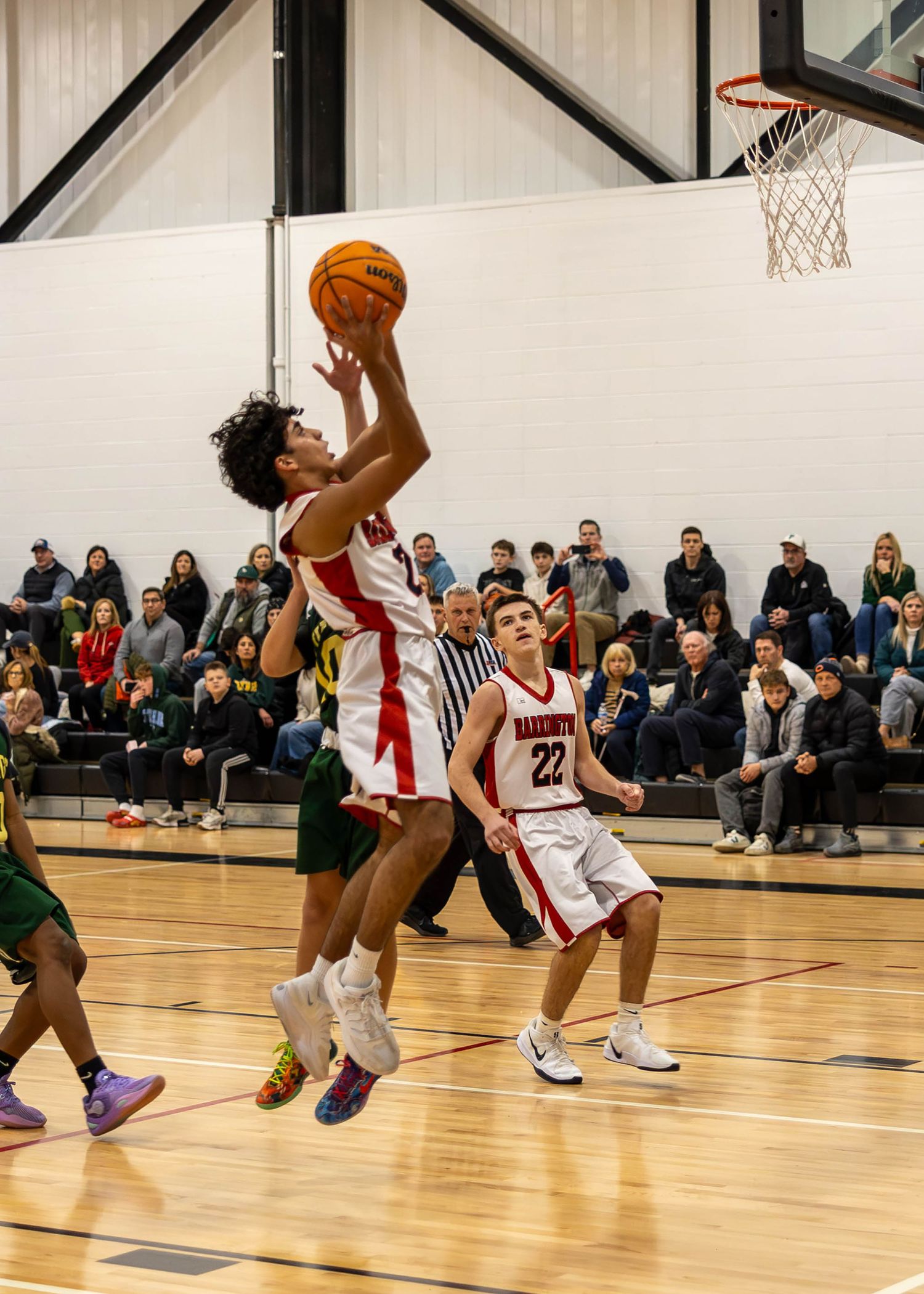 High school basketball player drives to the hoop during a competitive indoor game with teammates and crowd in background.