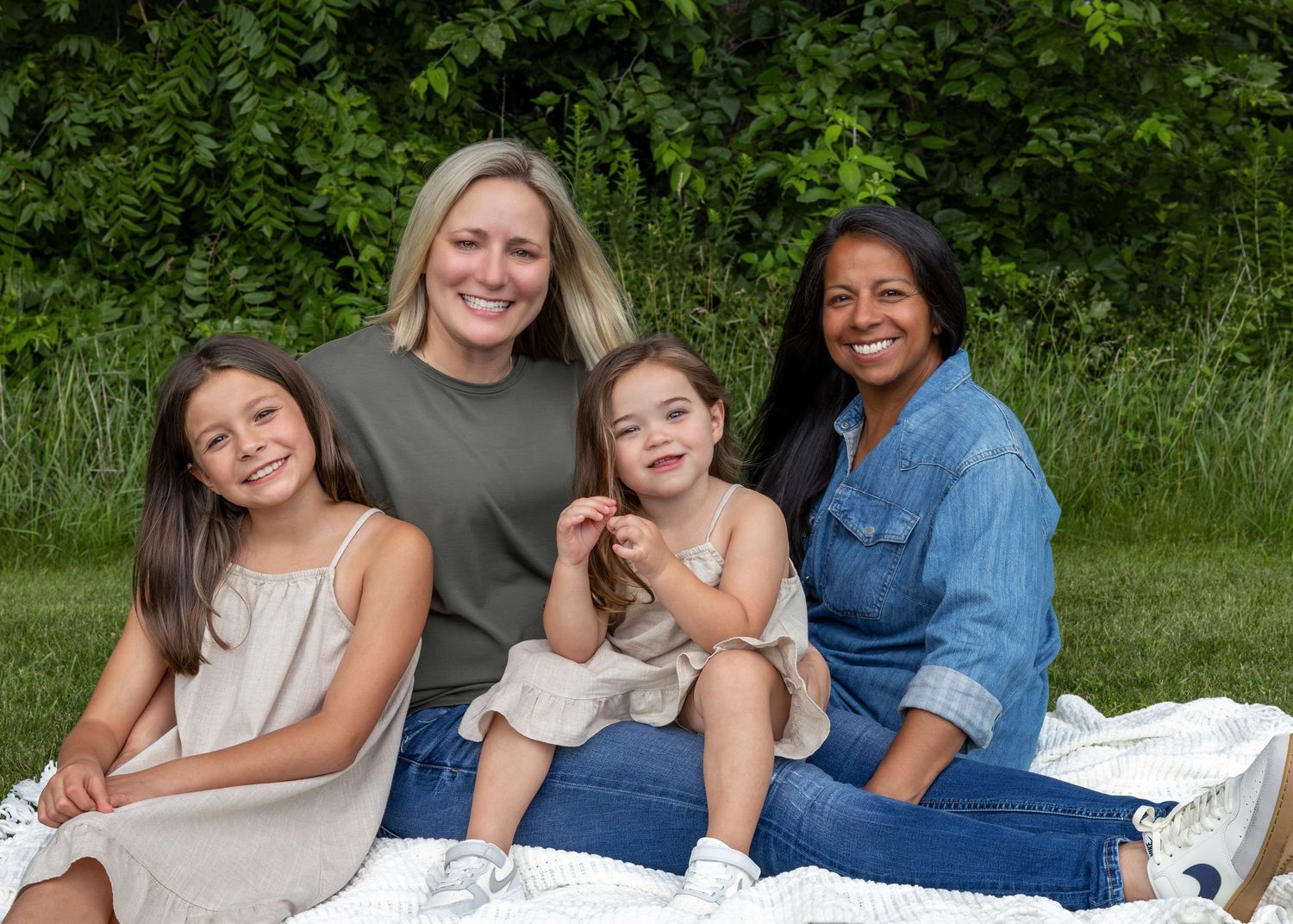 A family lounges casually together on a white blanket in an outdoor grassy setting during summer.