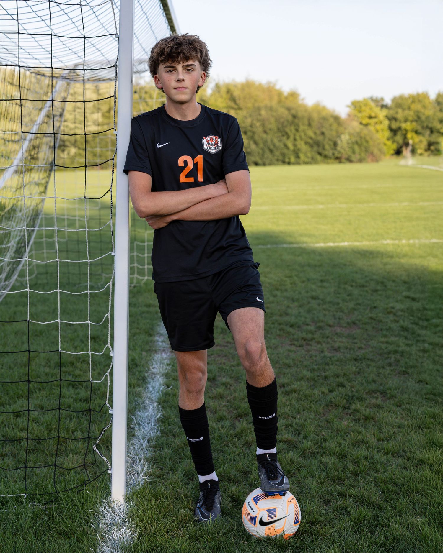 A soccer player in black uniform stands by a goal post on a sunny grass field with trees in the background.
