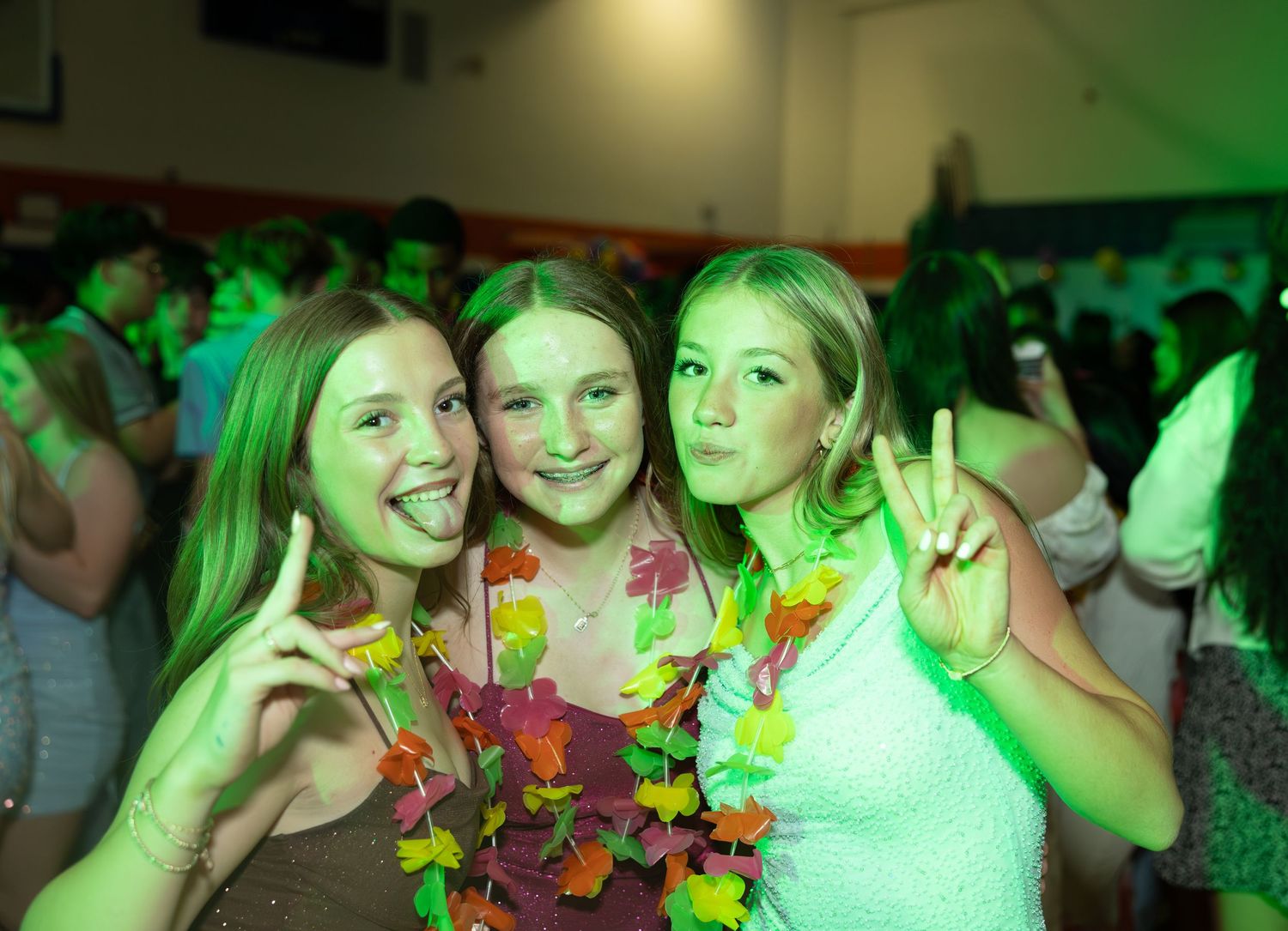 Three teenage girls posing together at a school dance, wearing colorful Hawaiian leis under green party lights.