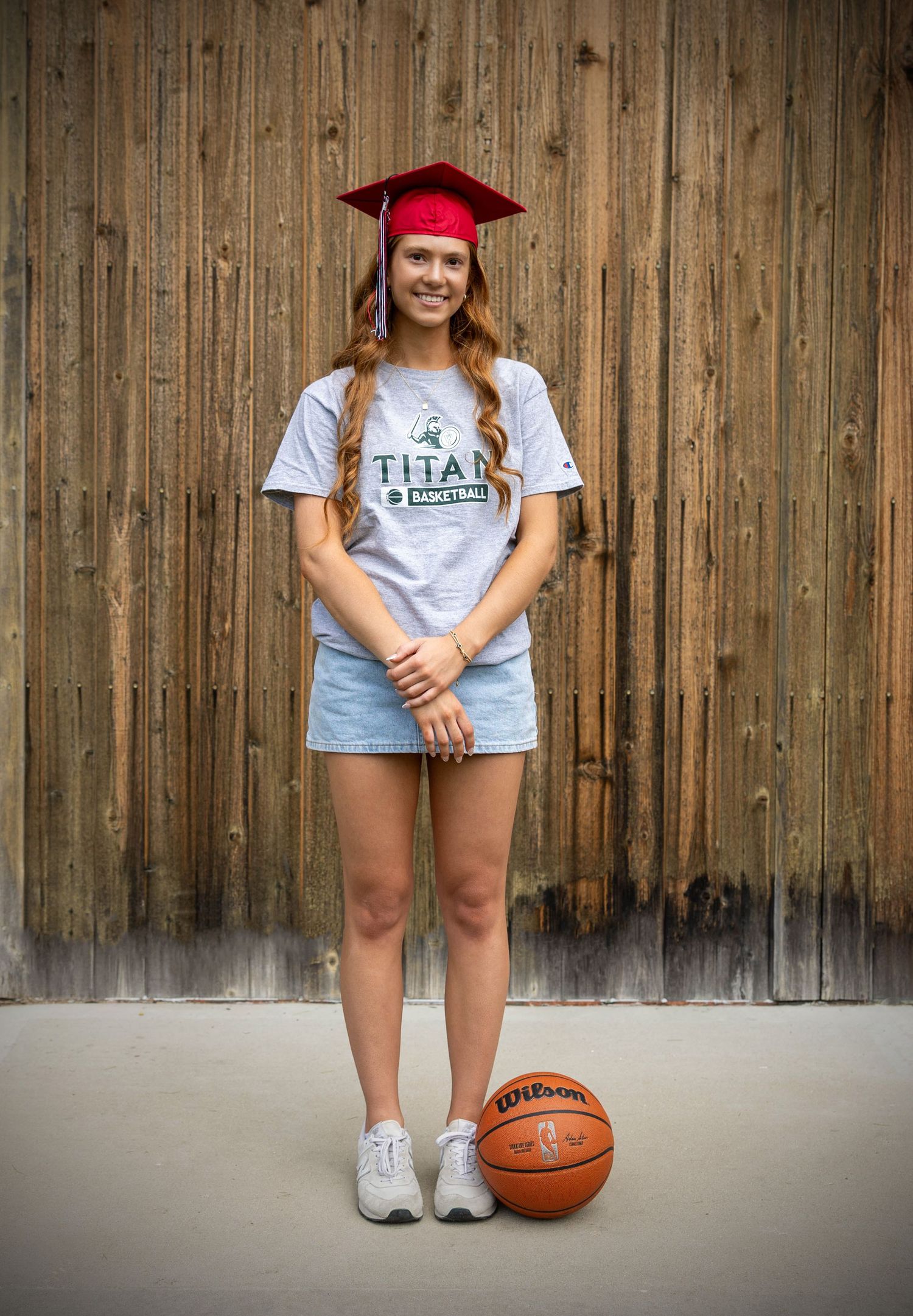 Graduate in red cap and Titan shirt poses with basketball against wooden wall background.
