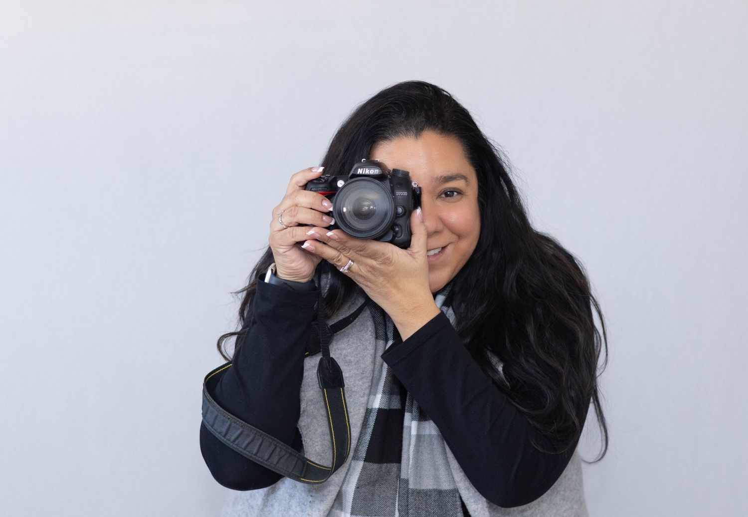 A photographer with long dark hair wearing a black sweater holds up a professional camera against a light gray background.