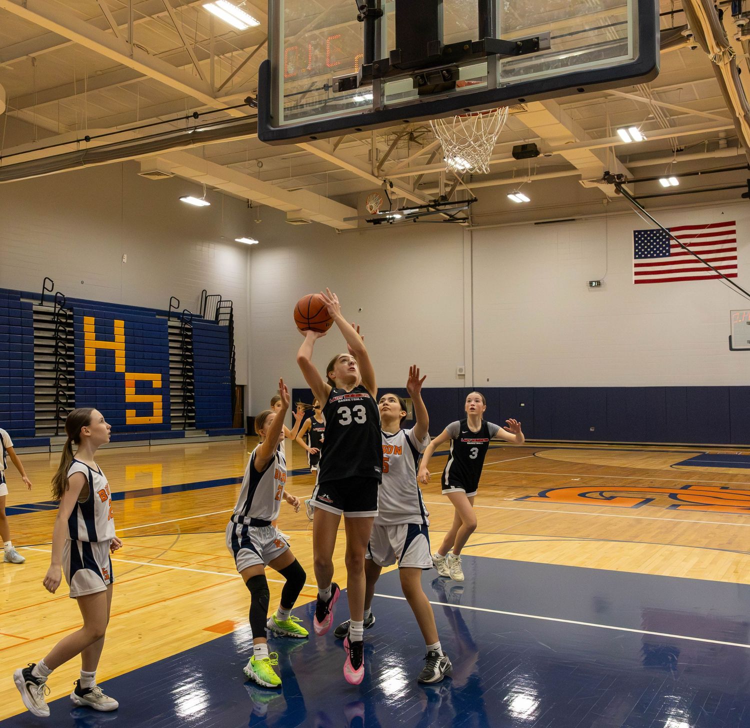 Girls basketball player #33 drives to the hoop during a high school gym game, teammates and opponents in pursuit.