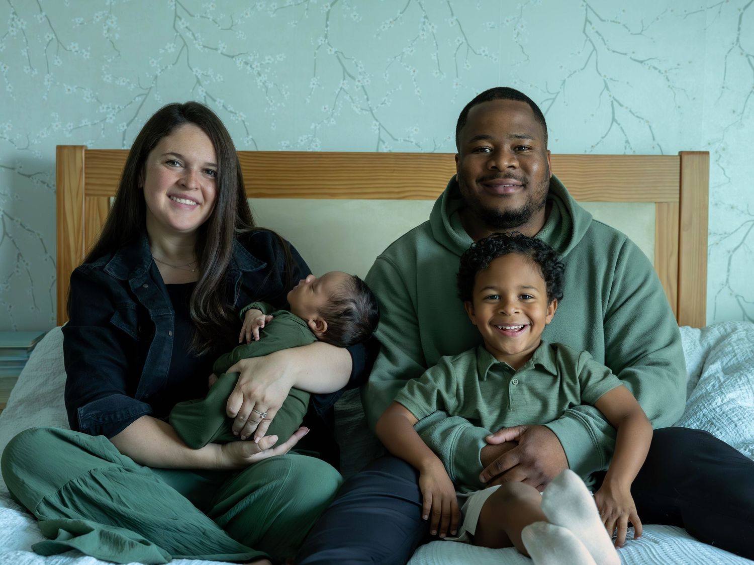 Happy interracial family posing on bed with newborn baby and young child in matching green outfits.
