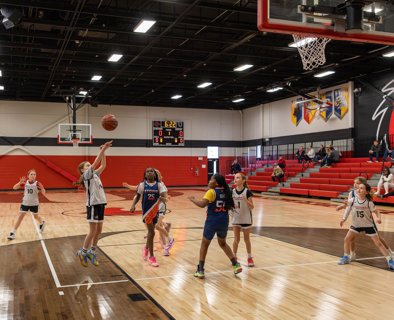 Girls youth basketball game in a red and white gymnasium, player shooting layup while teammates and opponents watch.