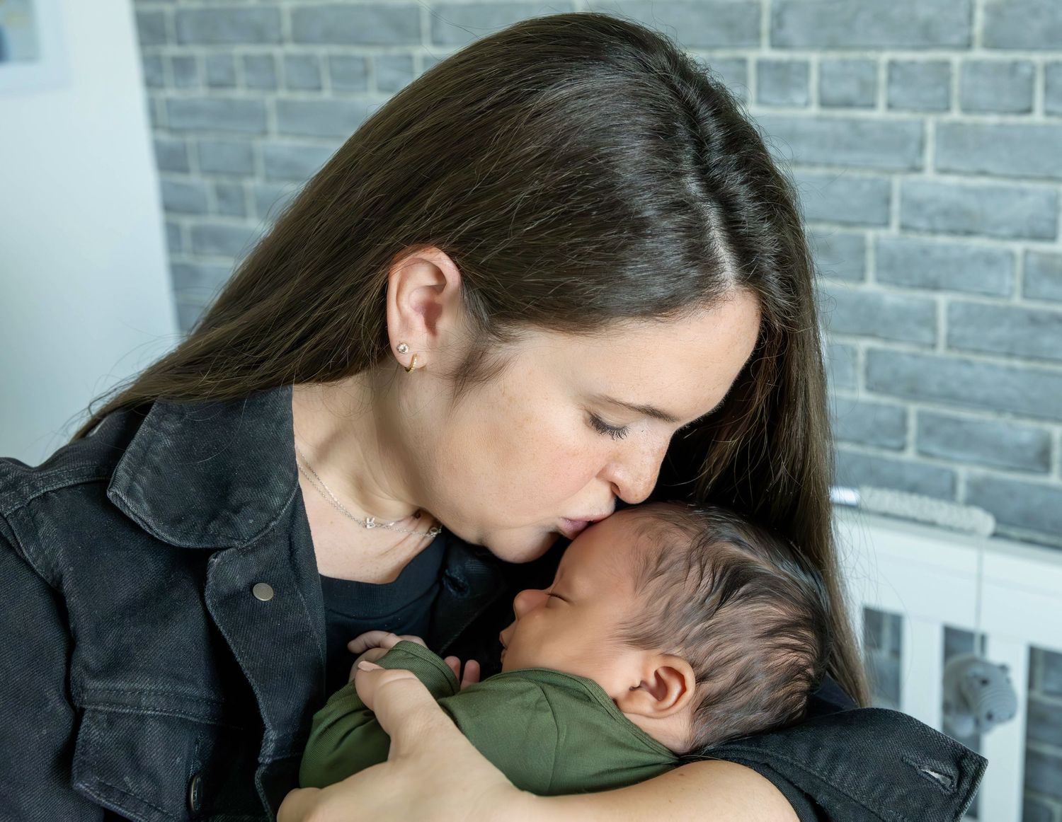 A mother tenderly kisses her newborn baby, cradling the infant in her arms against a brick wall backdrop.