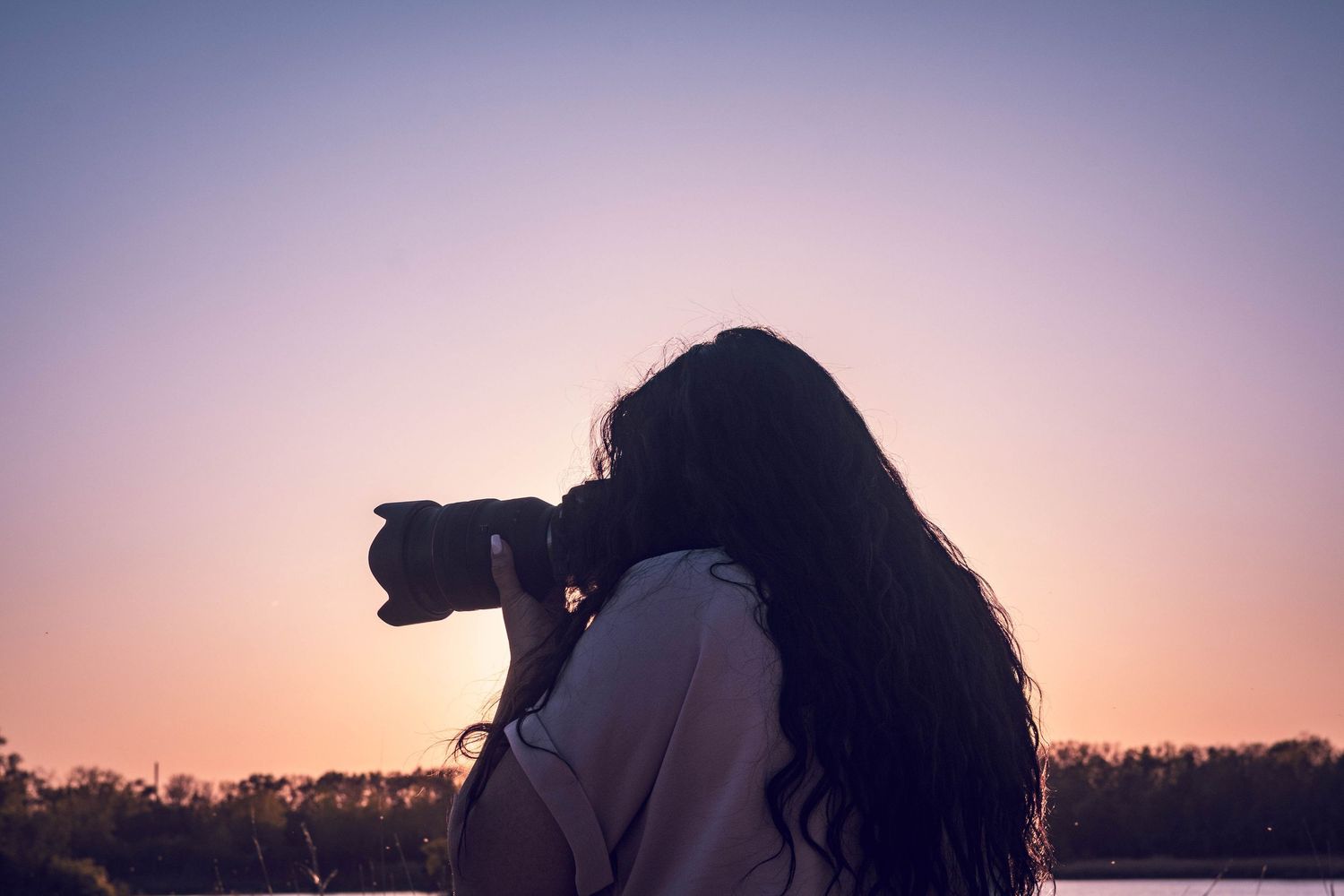 A silhouetted photographer with long hair taking photos during sunset with trees in the background.