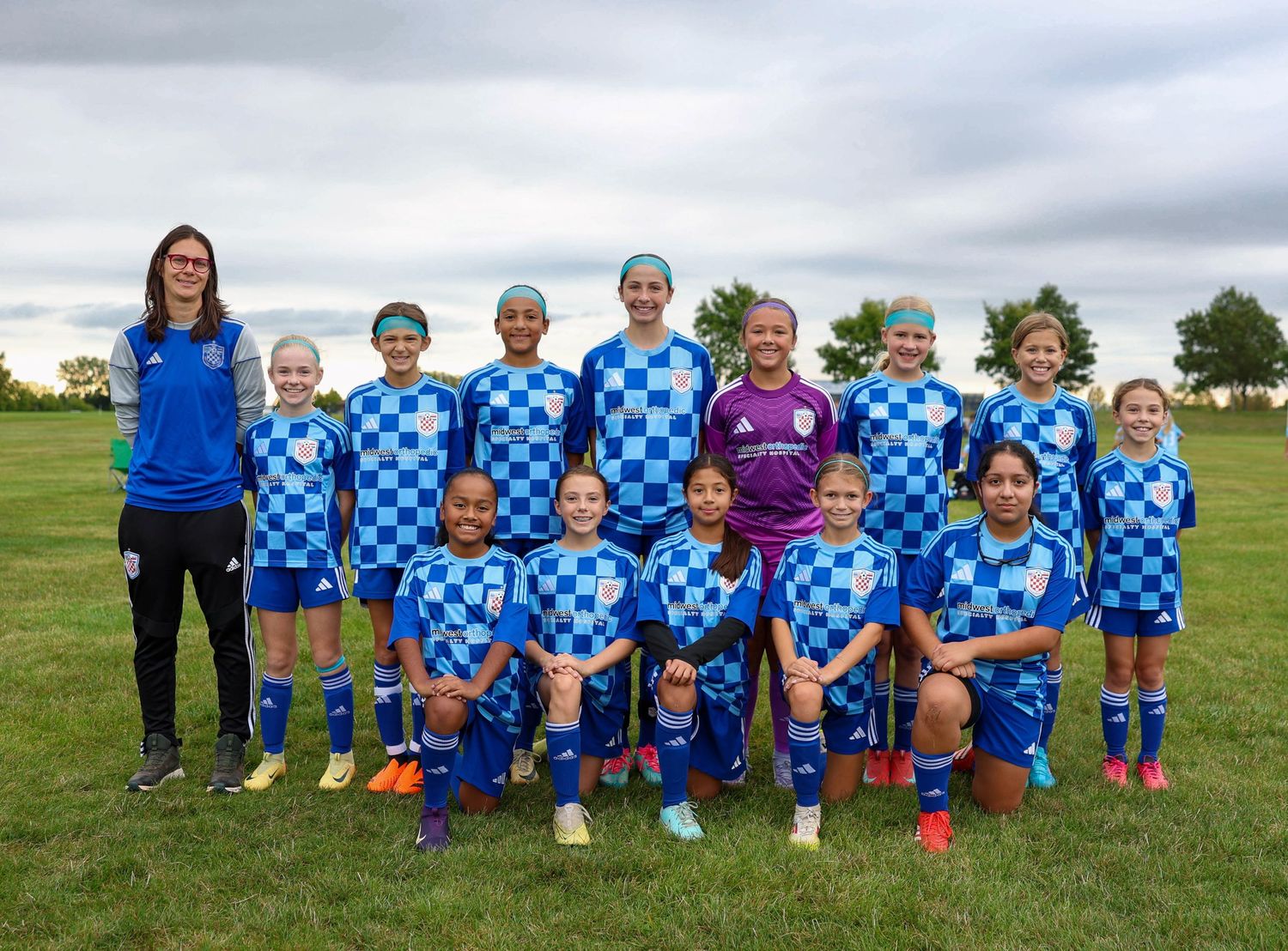 Youth soccer team wearing blue and white checkered jerseys poses together on a grass field with their coach.