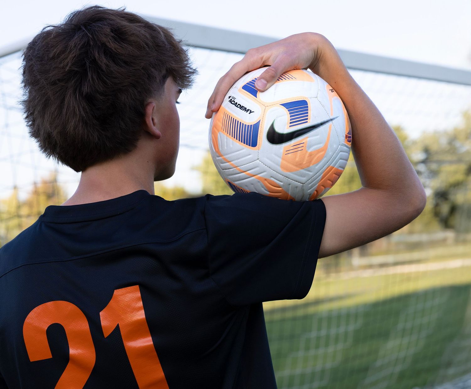 Soccer player wearing jersey number 21 holds Nike Premier League match ball on sunny field.