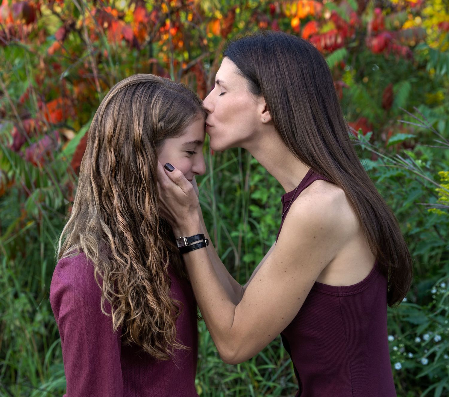 A tender moment shared between two people in burgundy tops against vibrant autumn foliage.