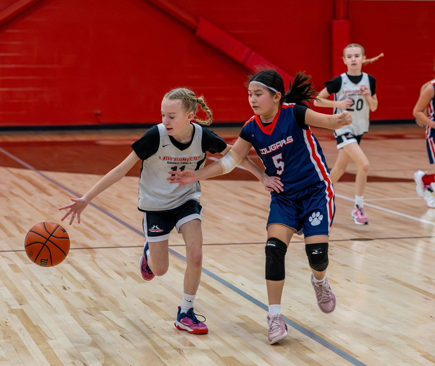 Two girls playing basketball in a gym, one dribbling past a defender wearing jersey number 5.