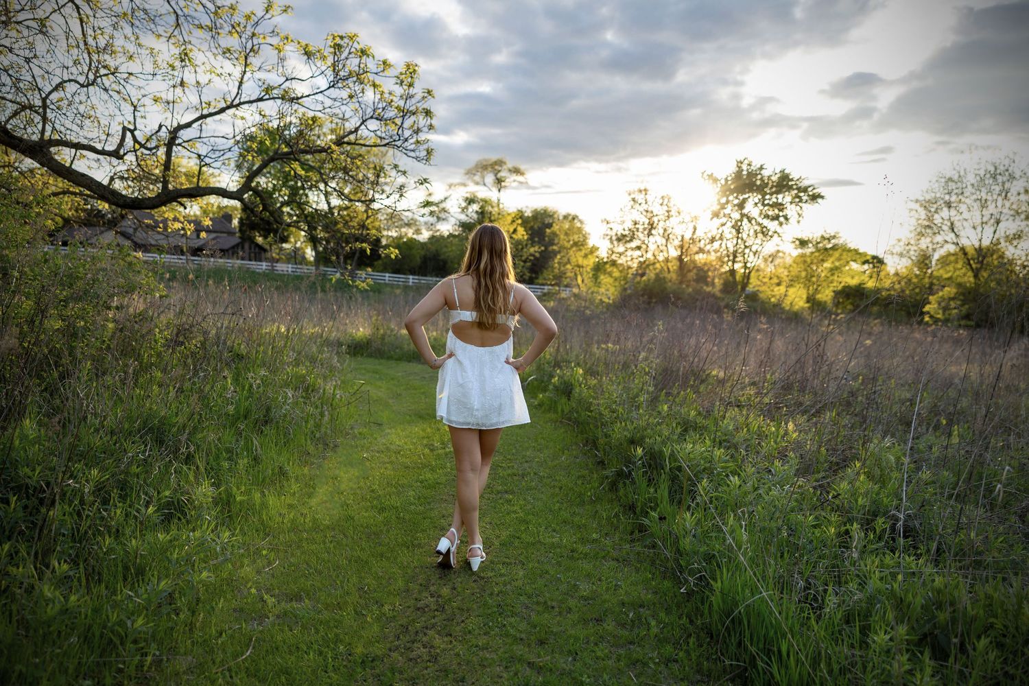 A person in a white dress takes a stroll along a grassy path at sunset in an overgrown meadow.