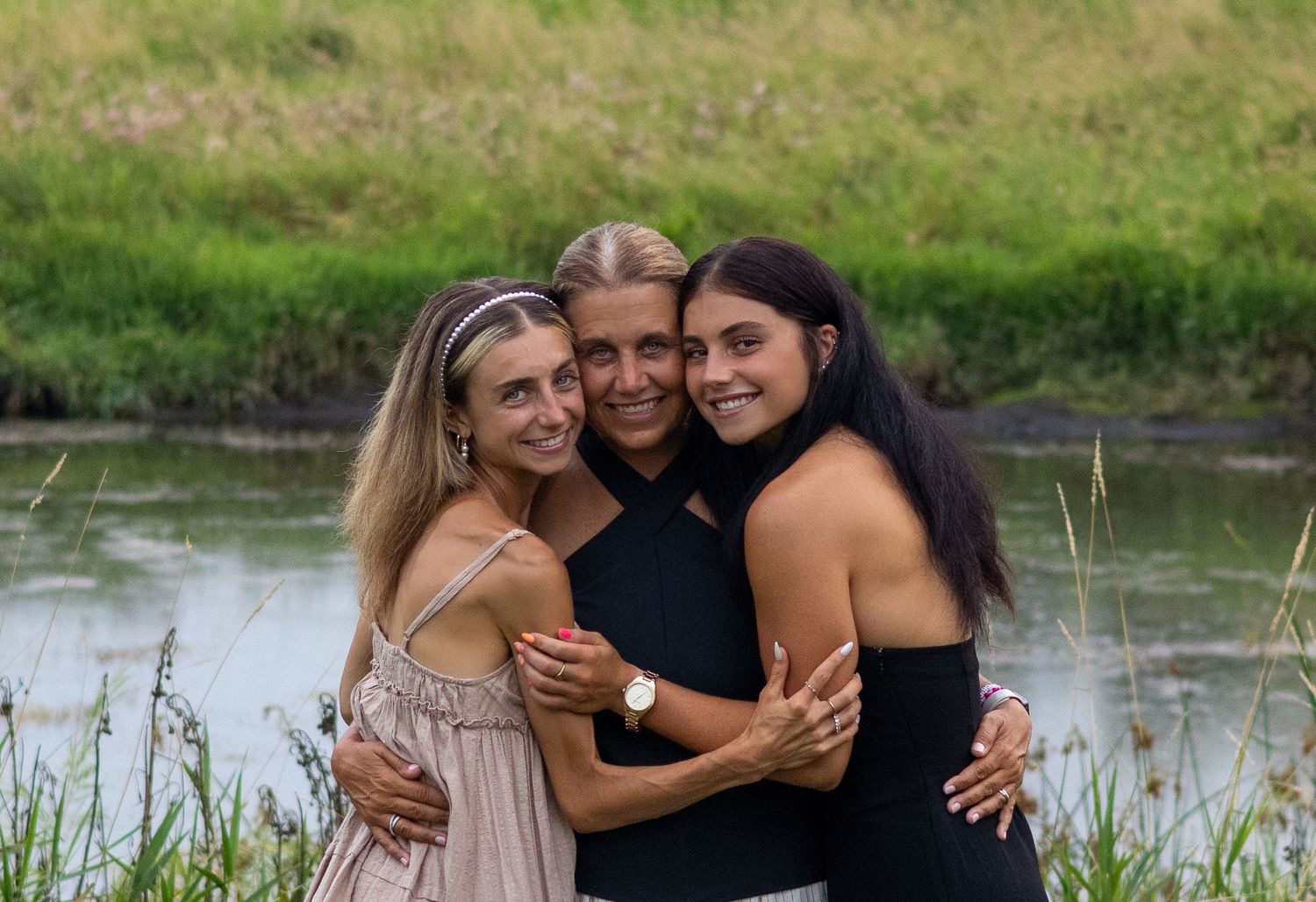 Three people embrace while posing together near a waterfront with grass and trees behind them.