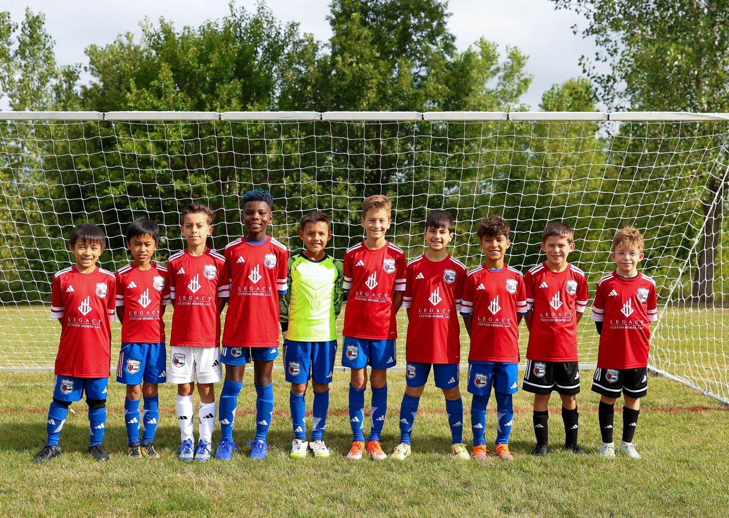 Youth soccer team in red jerseys and referees stand in front of a goal net on a sunny field for team photo.