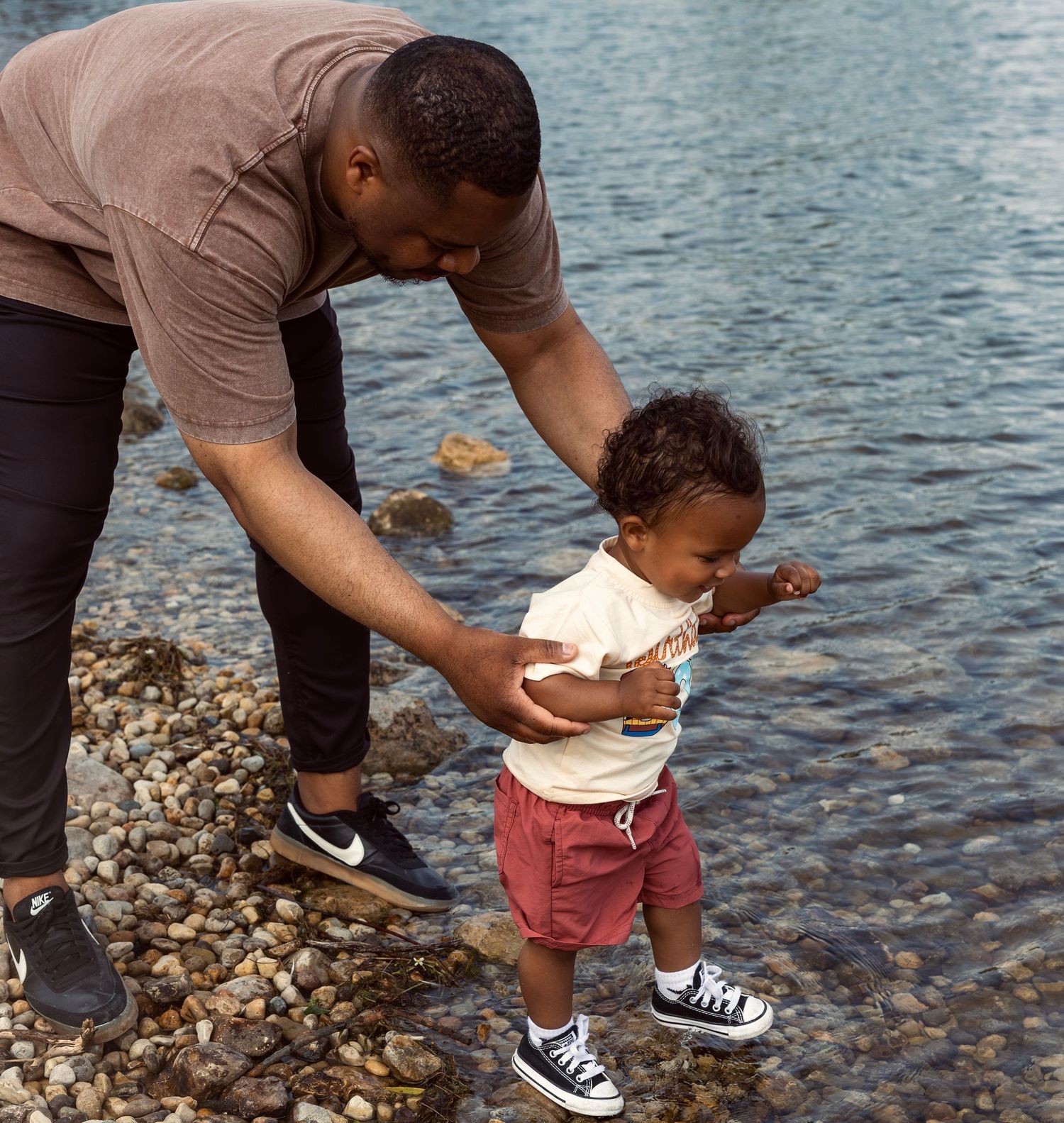 Father helping his toddler walk along a rocky shoreline near calm water on a sunny day.