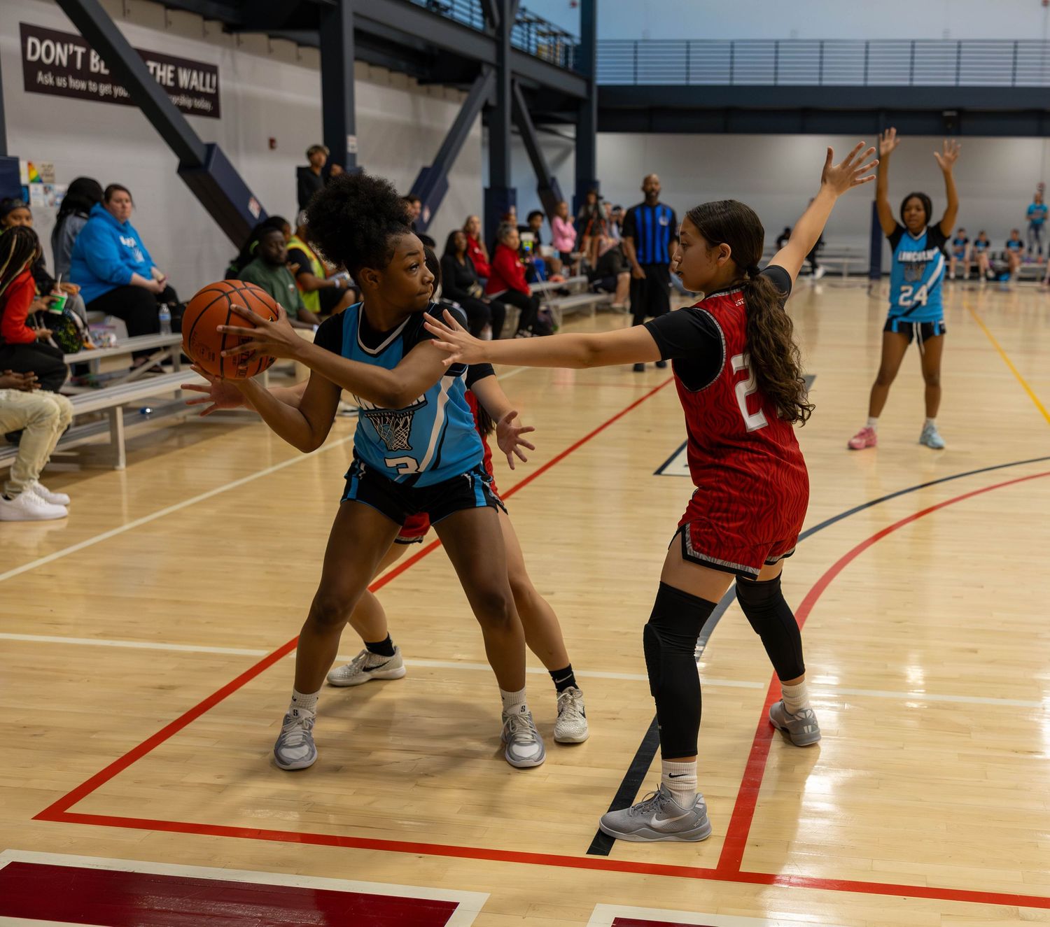 Two basketball players compete during an intense game on indoor court with spectators watching in background.