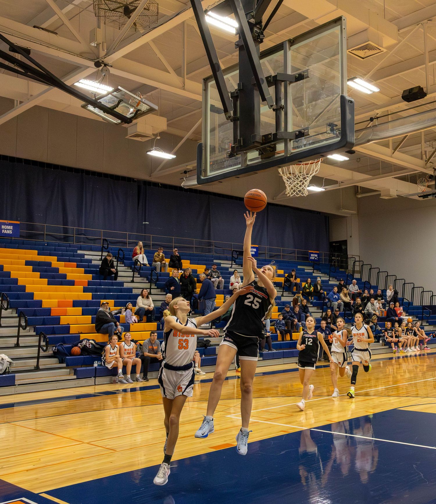 Player #25 in black executes a layup shot during a girls basketball game in a blue and gold gymnasium.