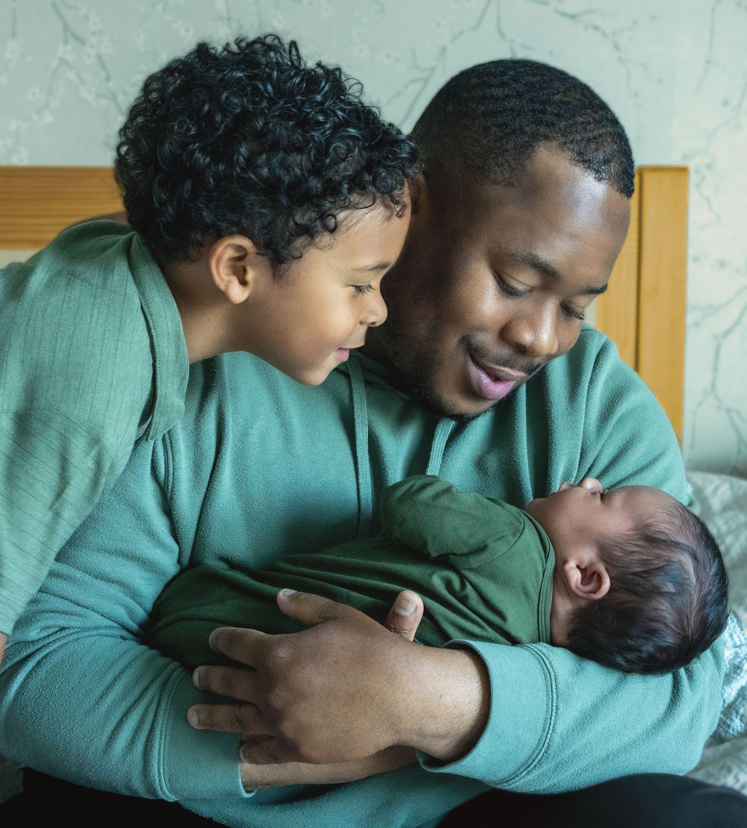 A father tenderly holds his newborn baby while an older child leans in affectionately, all wearing green.
