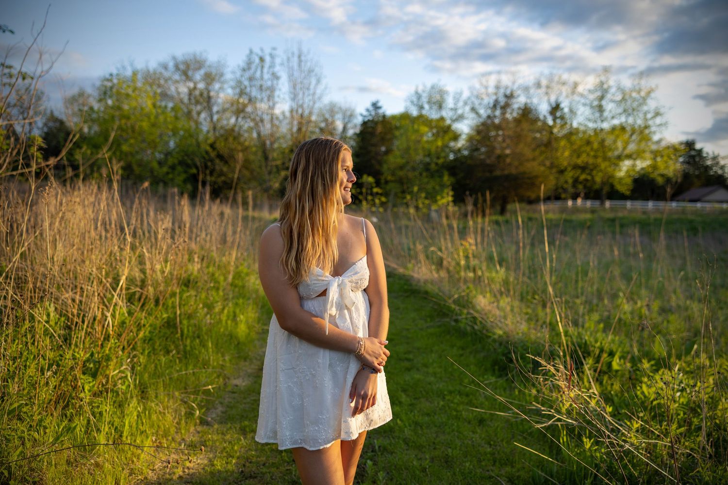 A person in a white sundress stands on a grass path during golden hour surrounded by tall meadow grass.
