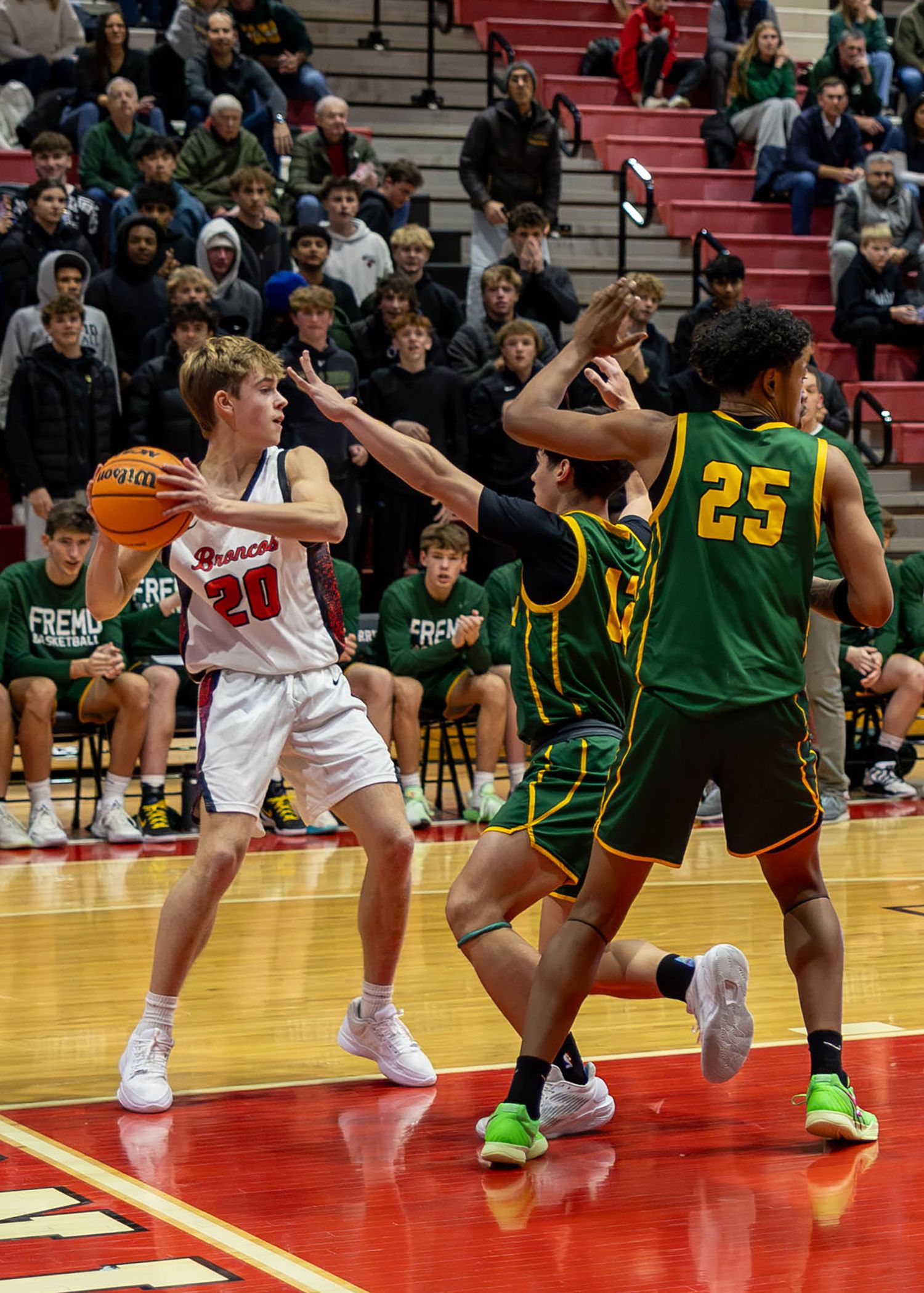 High school basketball player #20 in red uniform drives past Fremont defenders #25 during an indoor game.