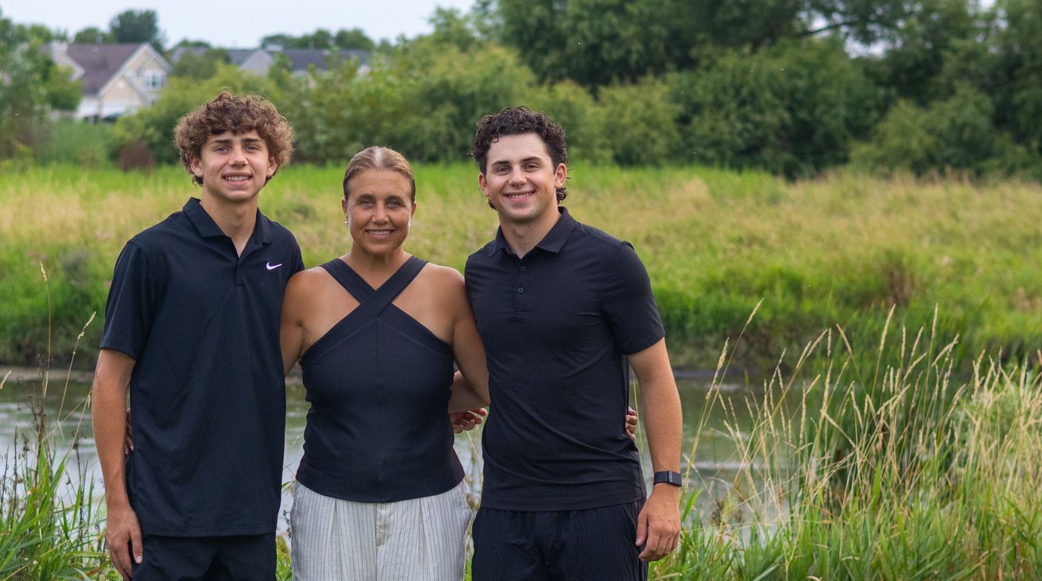 Three people smile while standing together outdoors near water and green foliage.