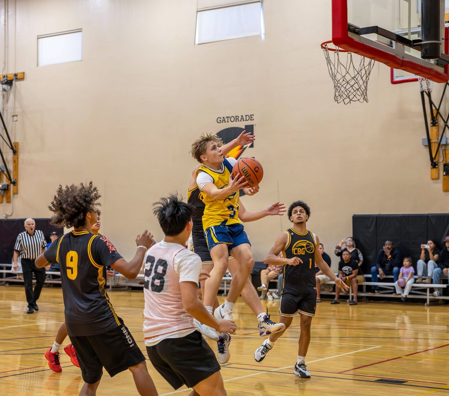 Youth basketball player drives to the hoop during an indoor game while defenders attempt to block the shot.
