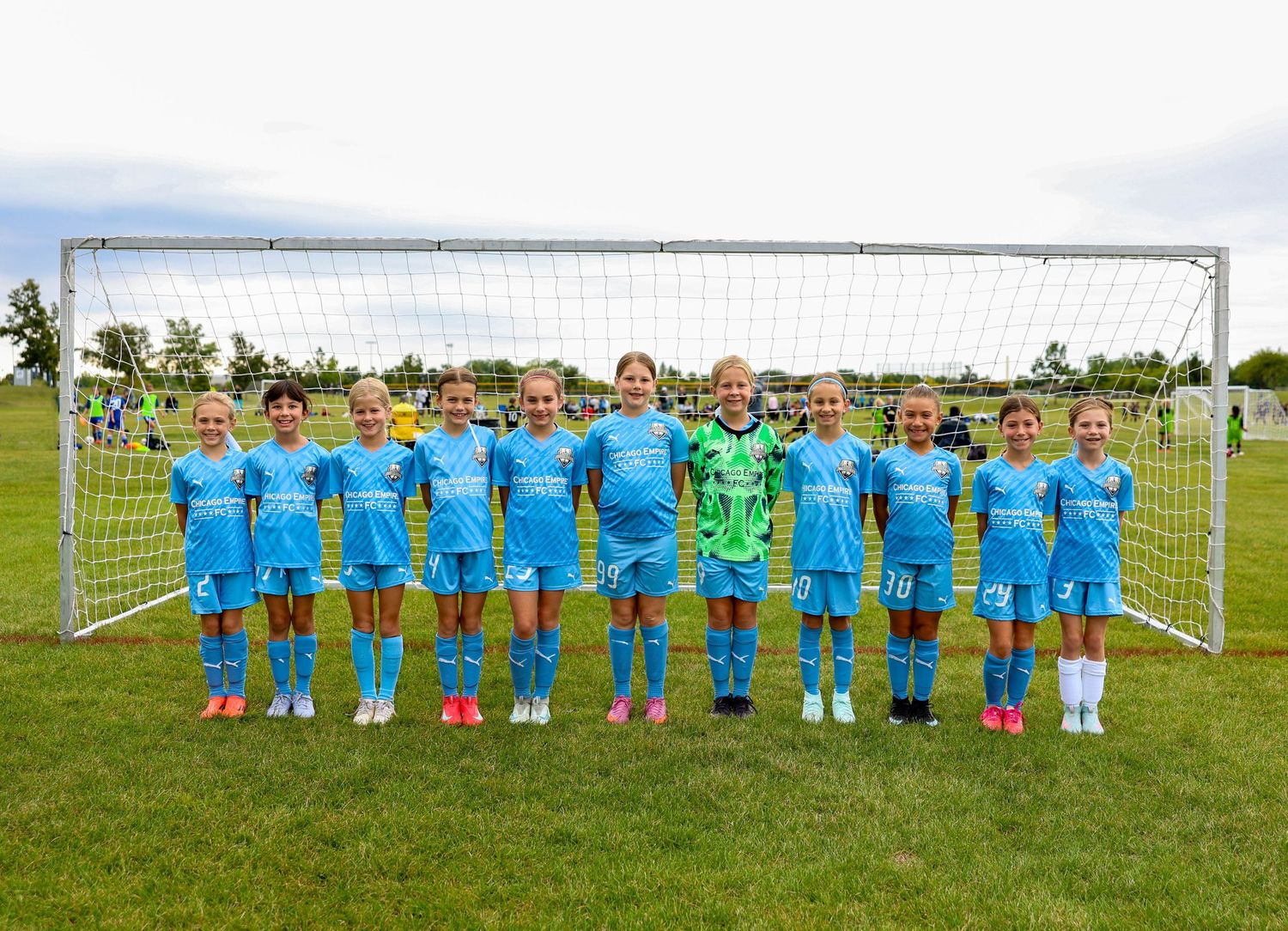 Youth soccer team in bright blue uniforms poses together on grass field with goal post in background.