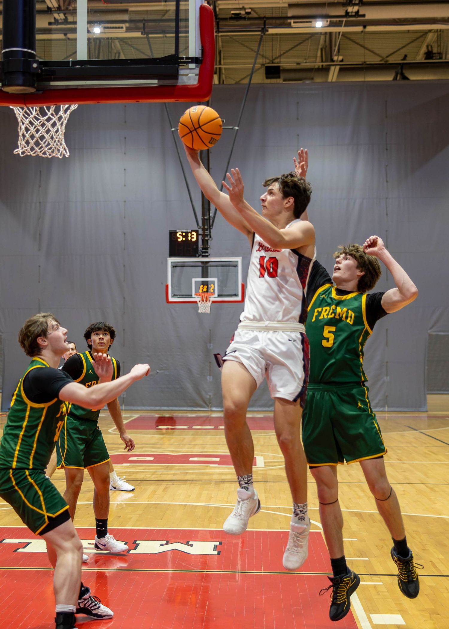 A high school basketball player in white jersey #10 leaps for a layup against green-uniformed defenders during an indoor game.