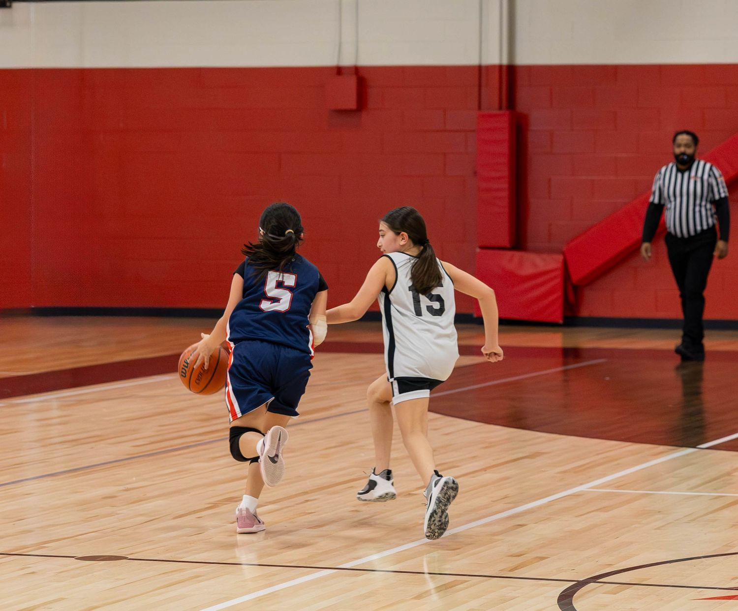 Two girls playing basketball in a gym, one dribbling in jersey number 5, referee watching in background.