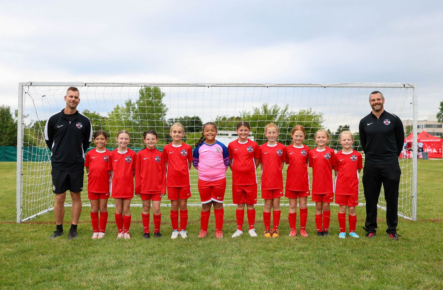 Youth soccer team in red uniforms stands in front of goal post with two coaches on grassy field.