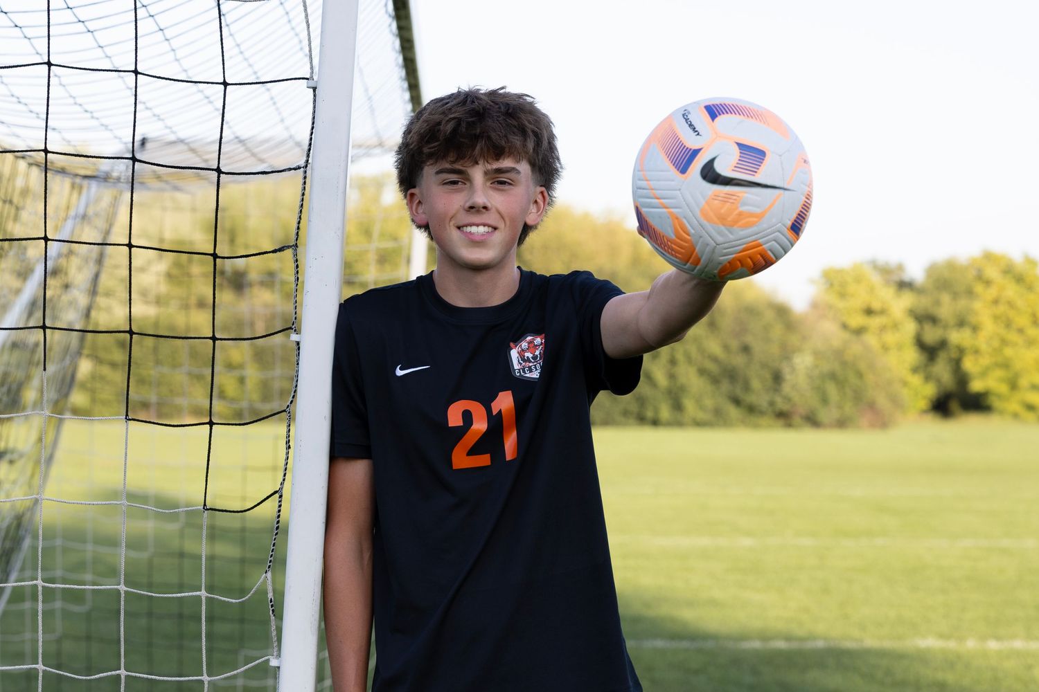 Soccer player in a black jersey smiles while standing next to a goal net on a sunny day.