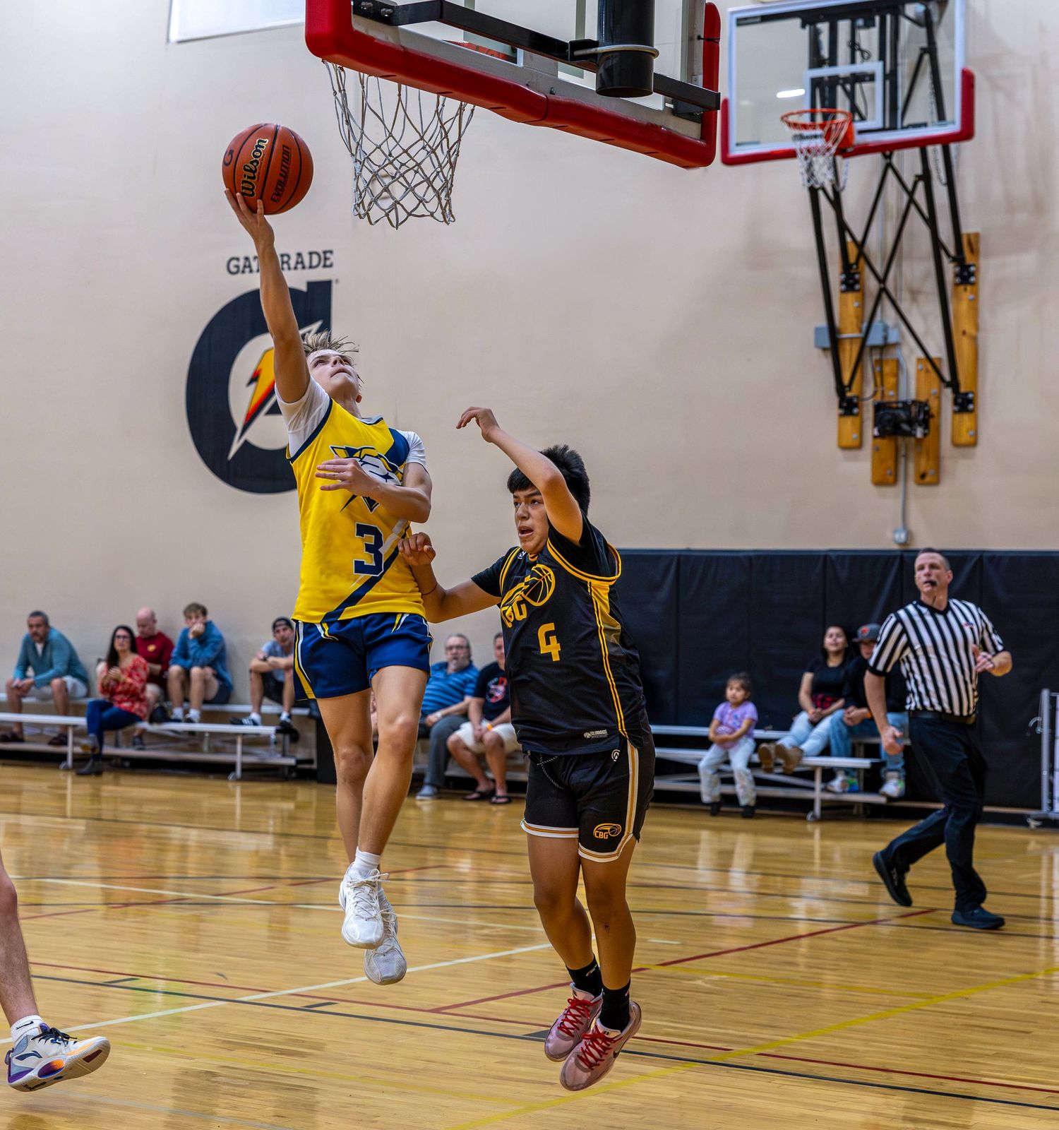 Youth basketball player in yellow jersey drives to the hoop past a defender during an indoor game.