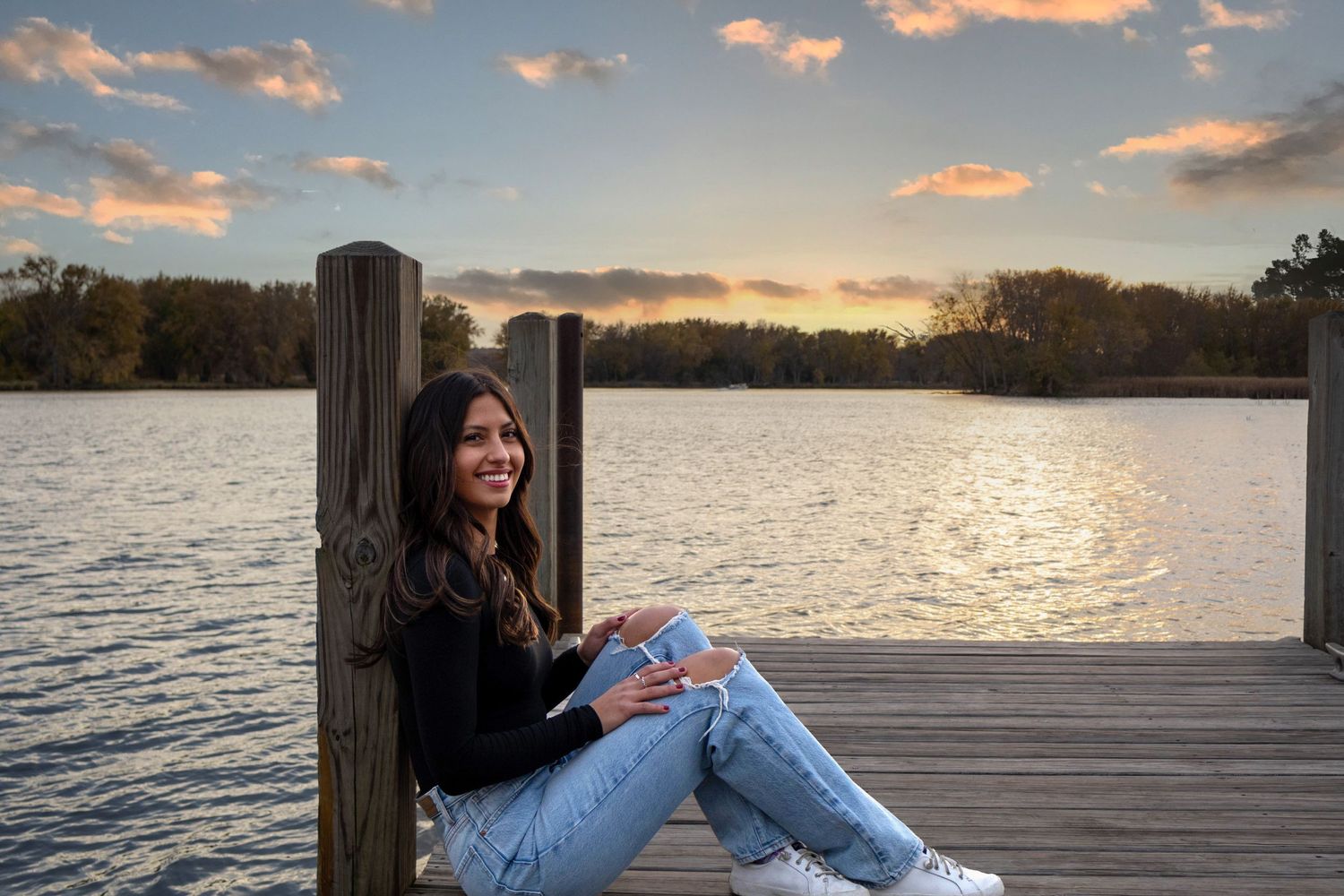senior student in black sweater and jeans sits on wooden dock during sunset over serene lake.