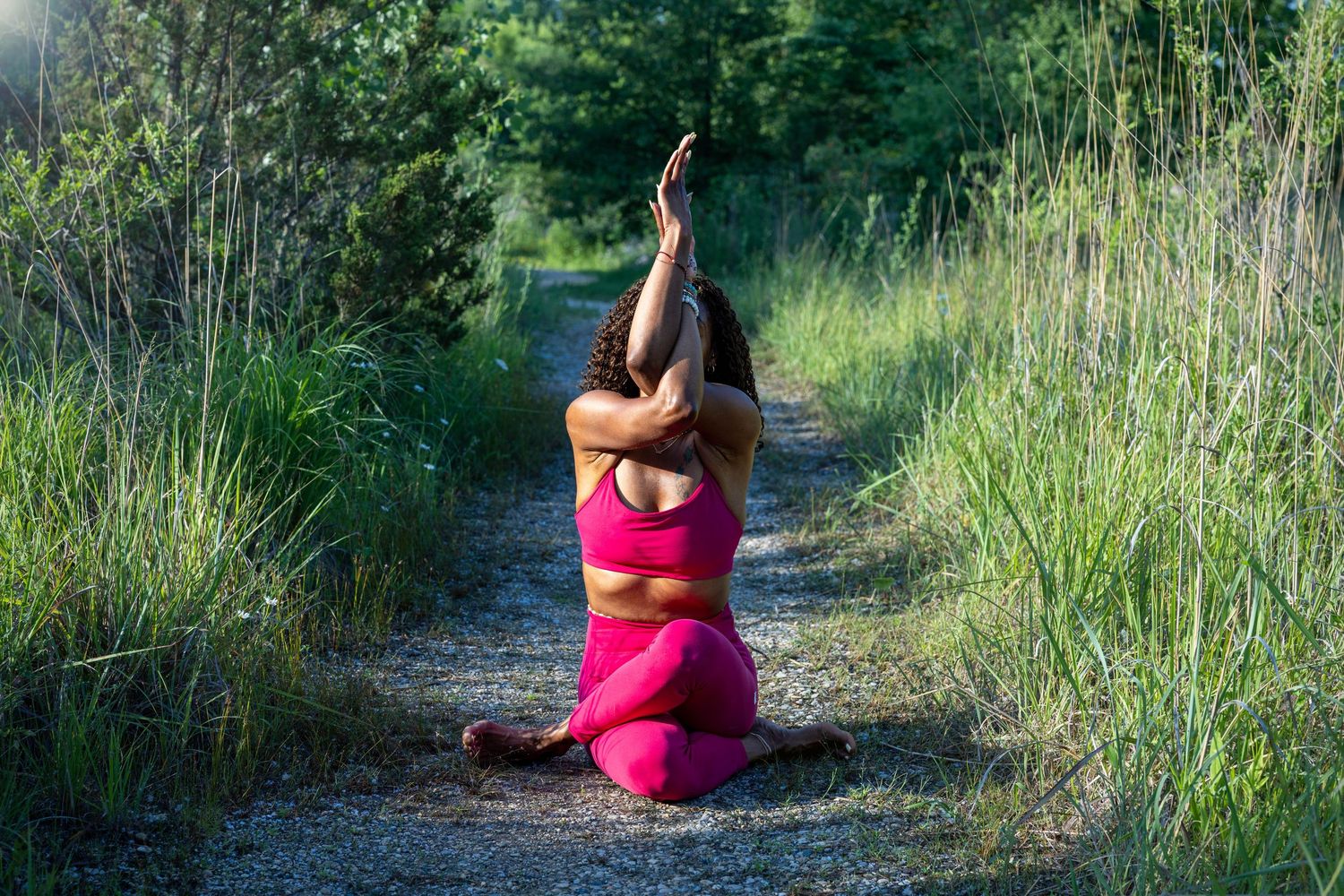 A figure in a hot pink outfit practices yoga poses on a grassy path surrounded by native vegetation.
