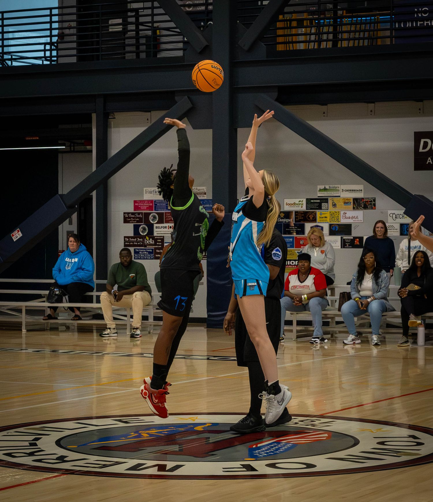 Basketball players jump high during a game tip-off with one in light blue and one in dark uniforms.