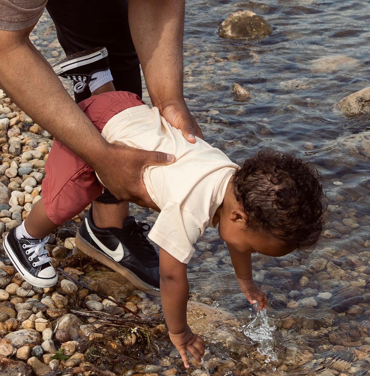 Adult holding toddler leaning toward rocky shoreline water, child touching the water with one hand.
