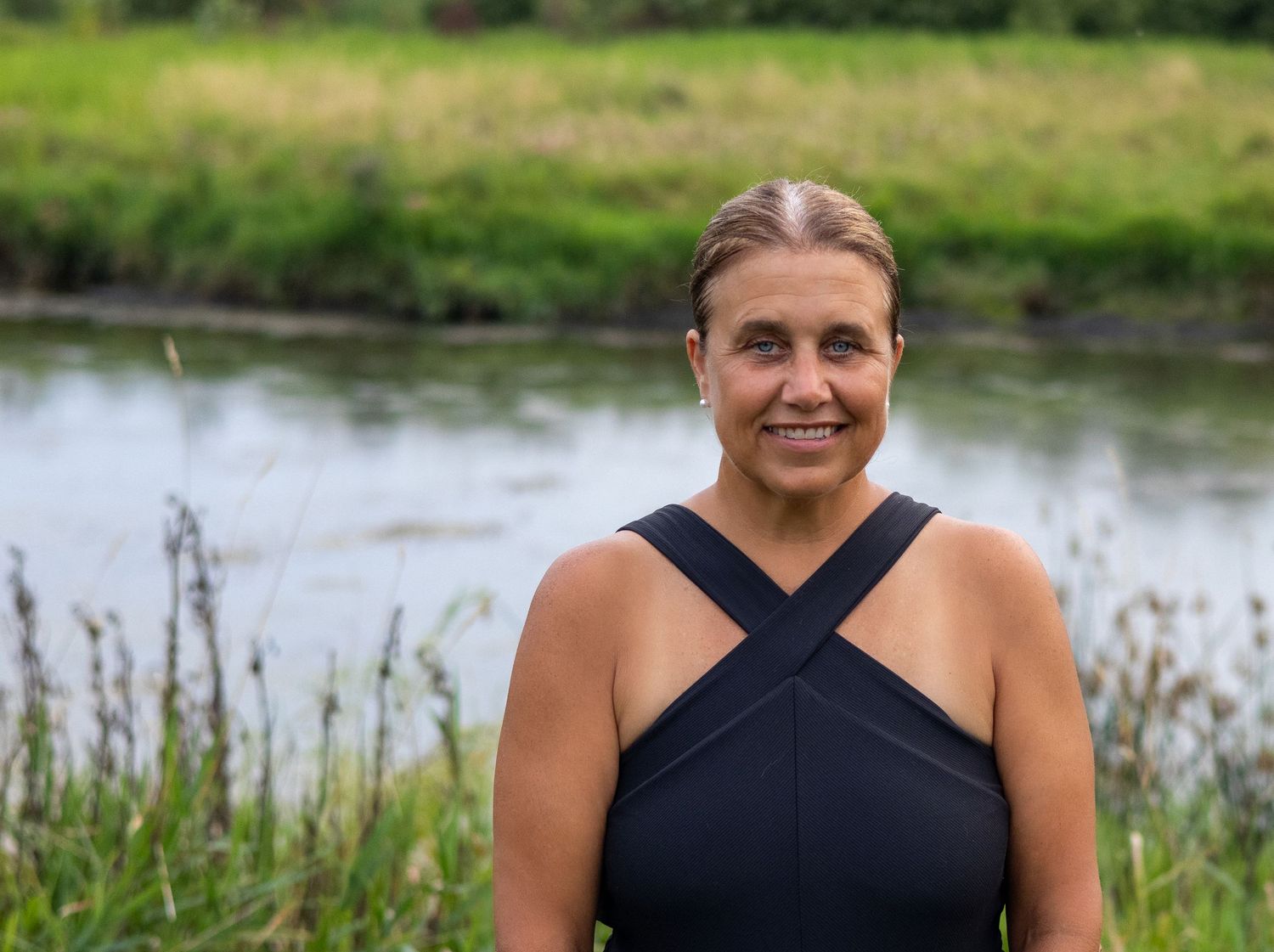 Mom in crossover top poses by pond with natural grass backdrop.