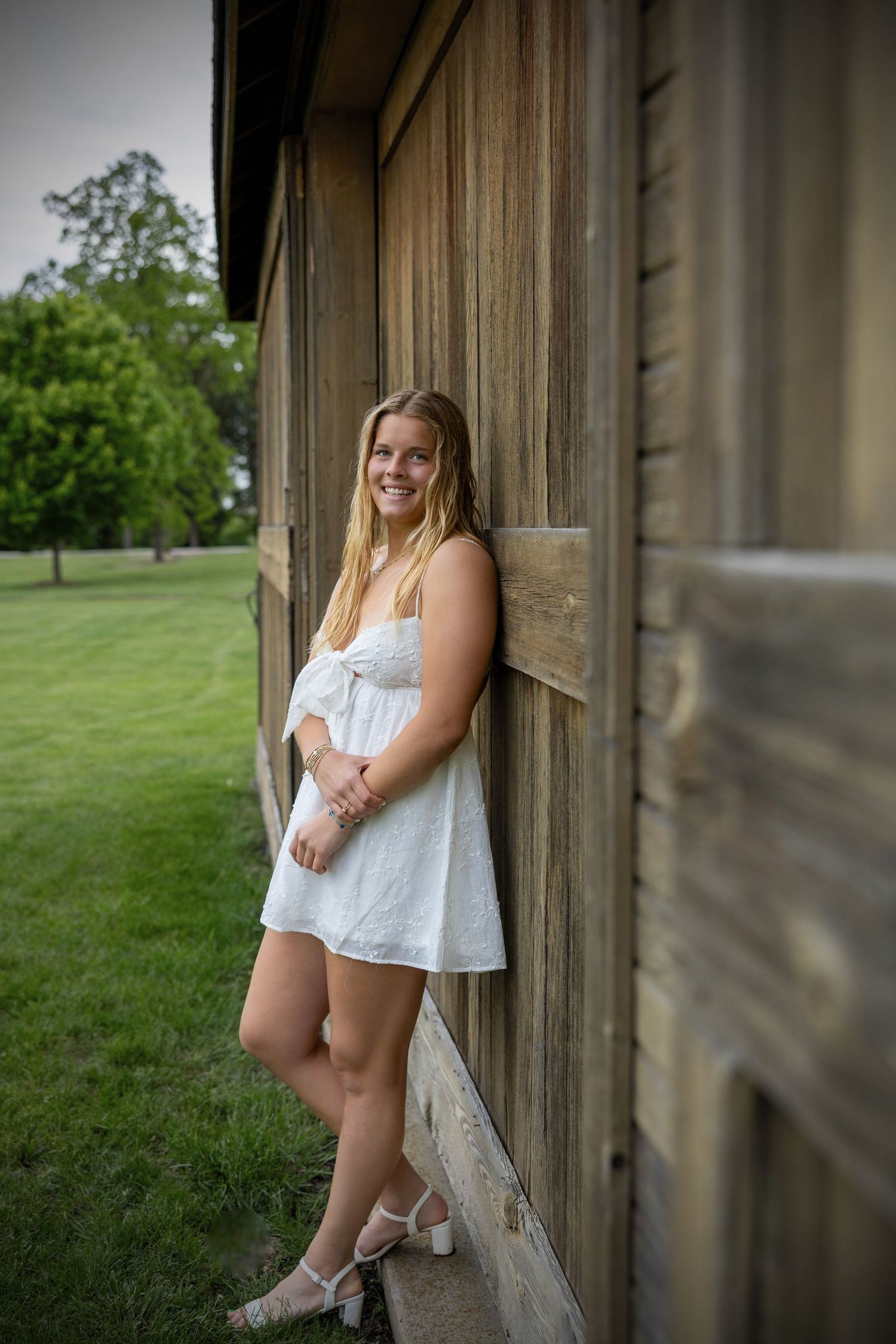 A person in a white summer dress poses against a rustic wooden barn wall on a sunny day with green grass in the background.