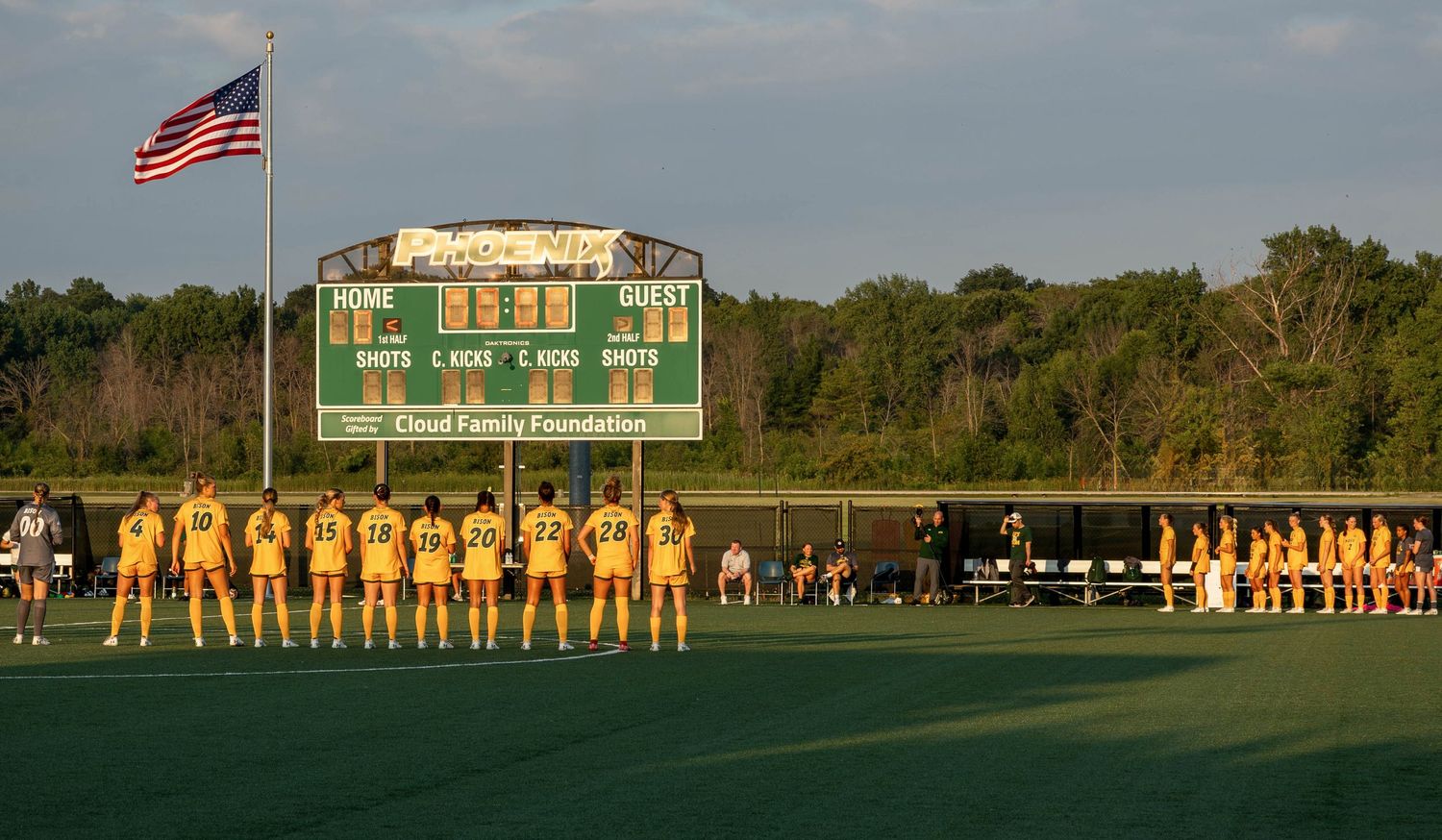 Soccer teams lined up on field before match with scoreboard and trees visible in background.
