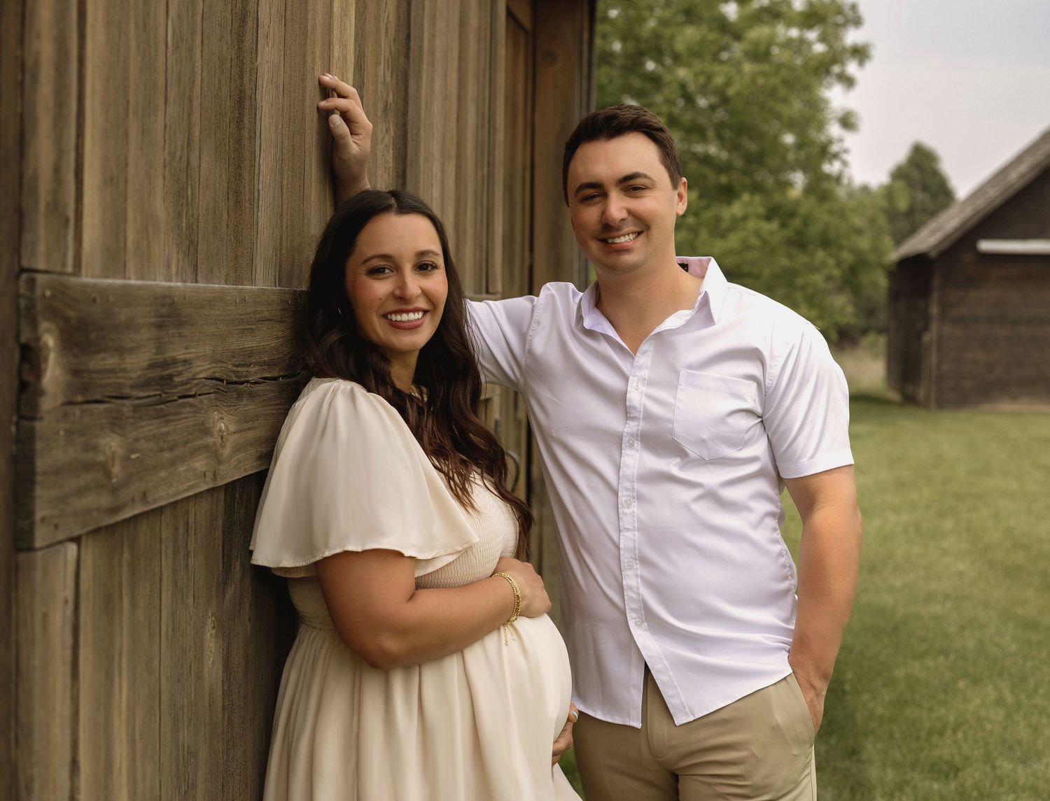 Smiling pregnant woman in white dress and man in light blue shirt pose together against rustic wooden barn wall outdoors.