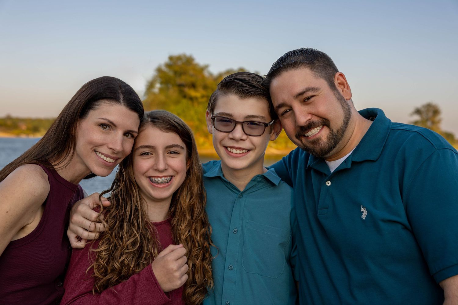 Happy family of four smiling outdoors near a lake at golden hour, wearing casual attire.