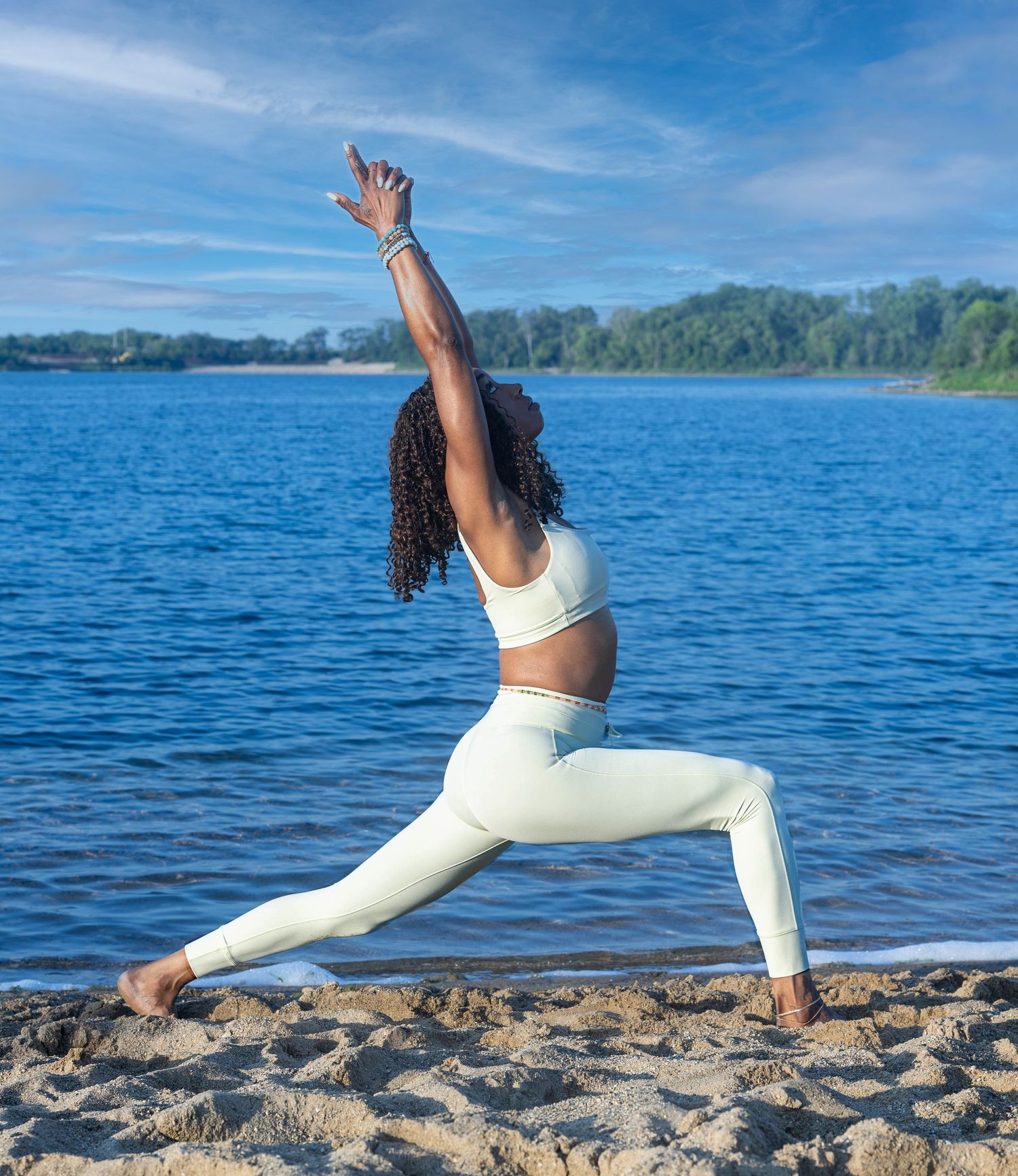 A woman practices a warrior yoga pose on a sandy beach by a lake, wearing white athletic wear on a sunny day.