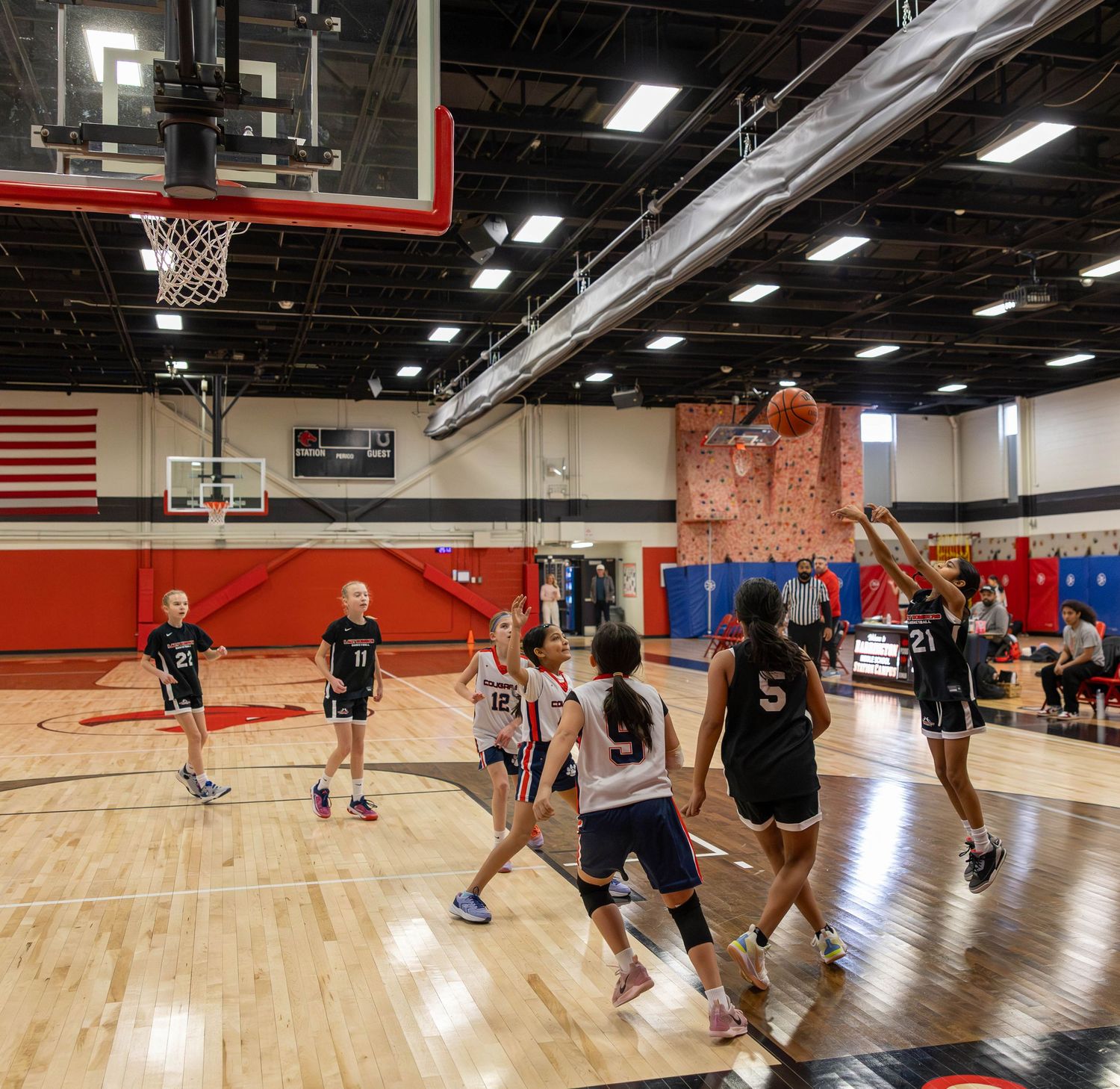 Youth girls basketball game in progress inside a modern indoor court with red walls and American flag.