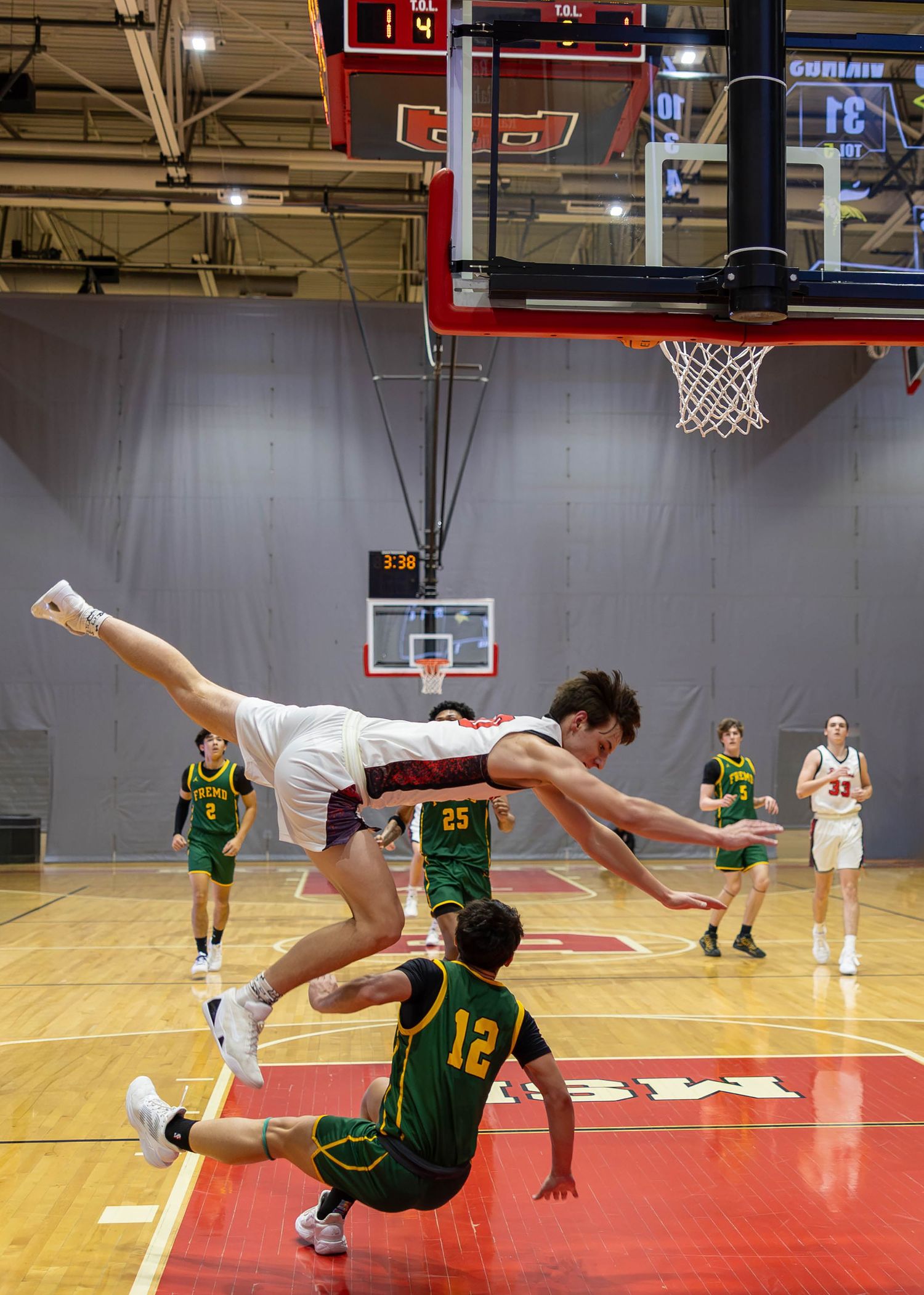 A basketball player in white dives over a fallen opponent #12 in green during an intense high school indoor basketball game.