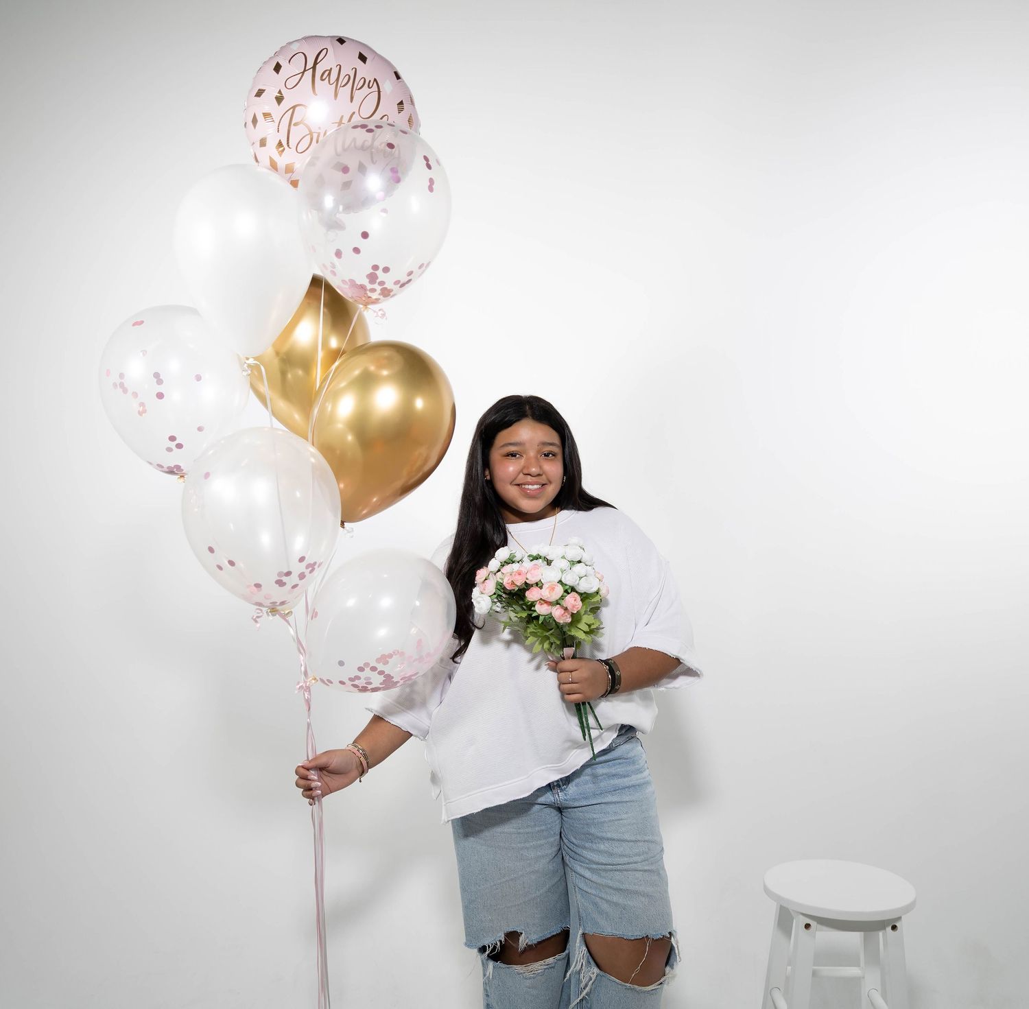 Young woman holding birthday balloons and pink flowers, smiling in white studio setting with gold and white balloon bouquet.