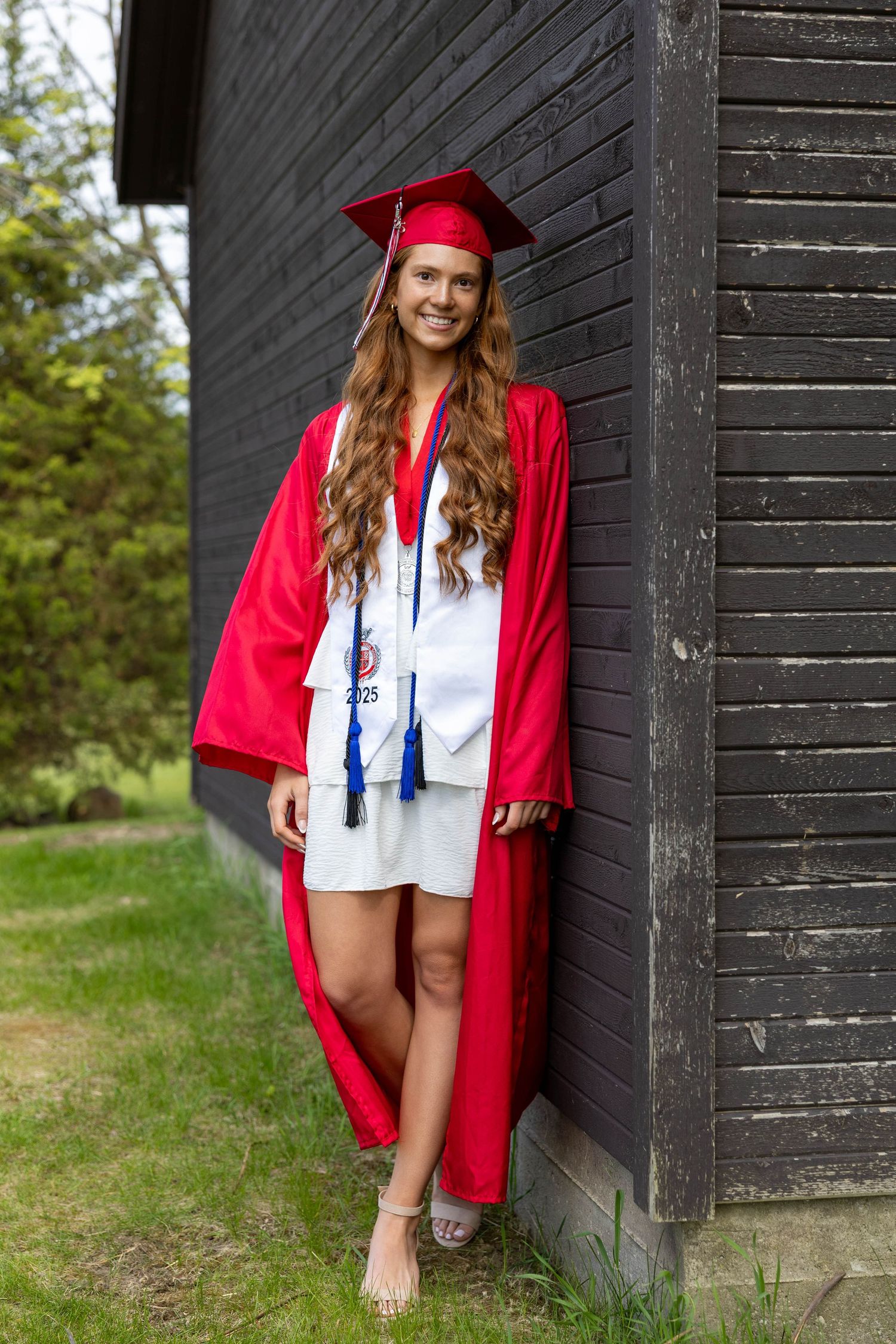 Young woman in red graduation cap and gown smiling outdoors against a dark wooden building.